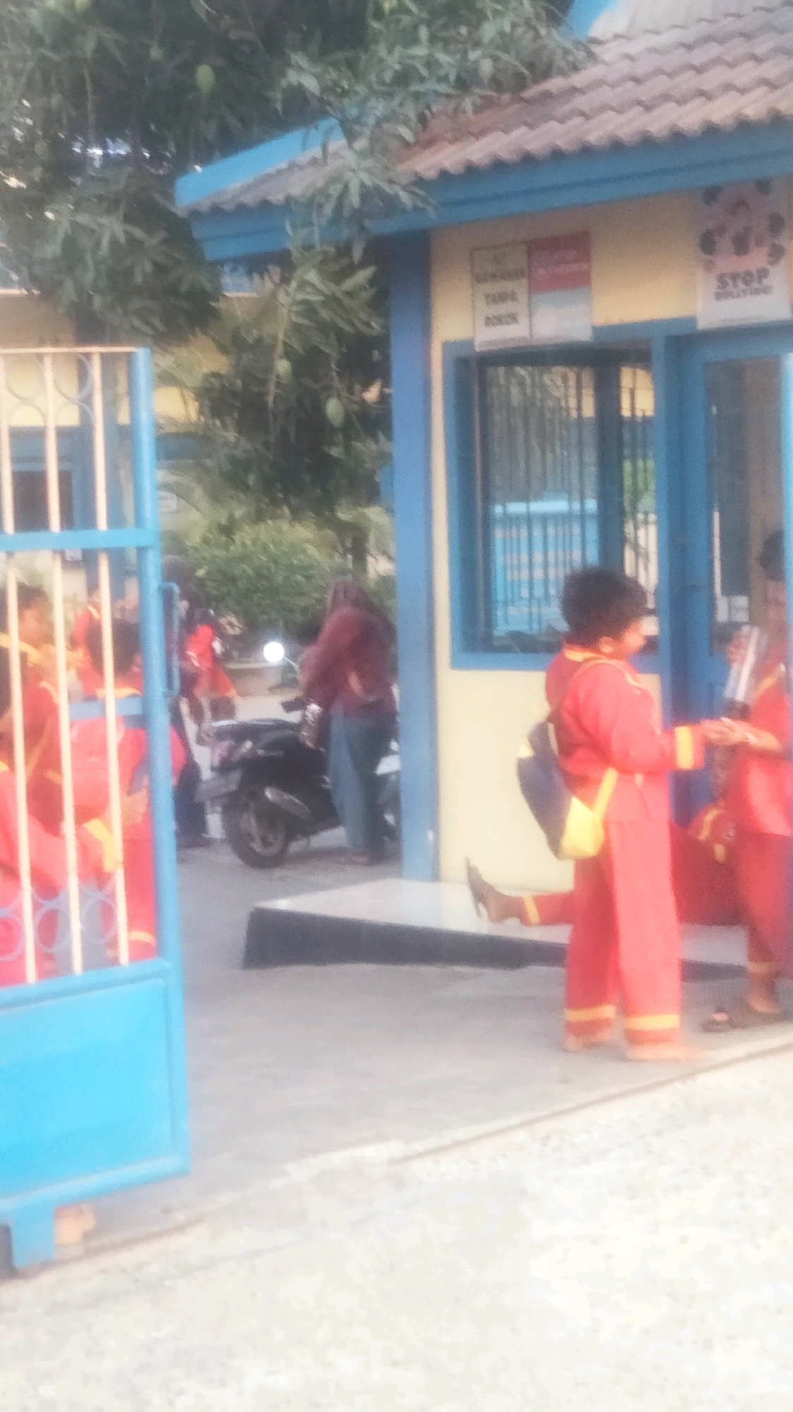 In front of a light yellow building with a blue roof trim and a brown shingled roof, a blue metal gate is partially open on the left side of the frame. Through the gate, several children in red uniforms with yellow trim are visible. A motorcycle is parked near the building. A woman in a dark red jacket and blue jeans stands near the motorcycle. To the right of the building entrance, a person in a red uniform with a yellow bag over their shoulder is walking away from the building. Another person in a similar red uniform is to their right, also facing away. There are signs on the building's wall, one appears to be a "STOP BULLYING" sign with a graphic of hands. Trees with green leaves and some green fruit are visible behind the building and to the left. The ground is paved. The location is identified as Palang, Indonesia.
