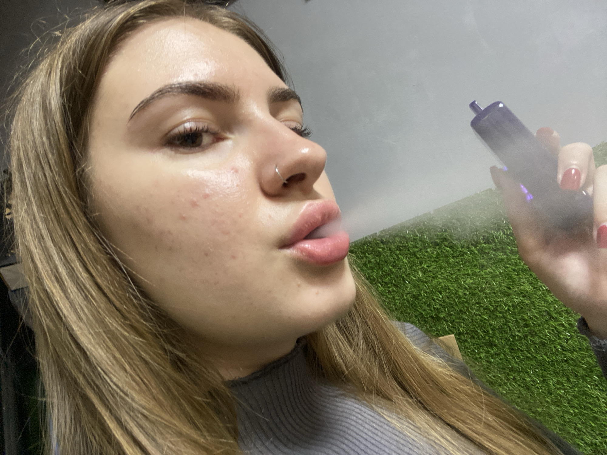 A close-up, medium-shot photograph shows a young, fair-skinned woman with long, blonde hair, positioned in the foreground. She is holding a purple vape pen in her right hand, from which a plume of white vapor is emanating towards her face. Her lips are slightly parted as she exhales the vapor, and she wears a silver nose ring. The background features a wall covered in artificial green turf. The lighting is diffused, casting a soft glow on the woman's face, and the overall atmosphere is casual. The image does not contain any discernible elements that would place it specifically in the city of Дьяково, Russia.