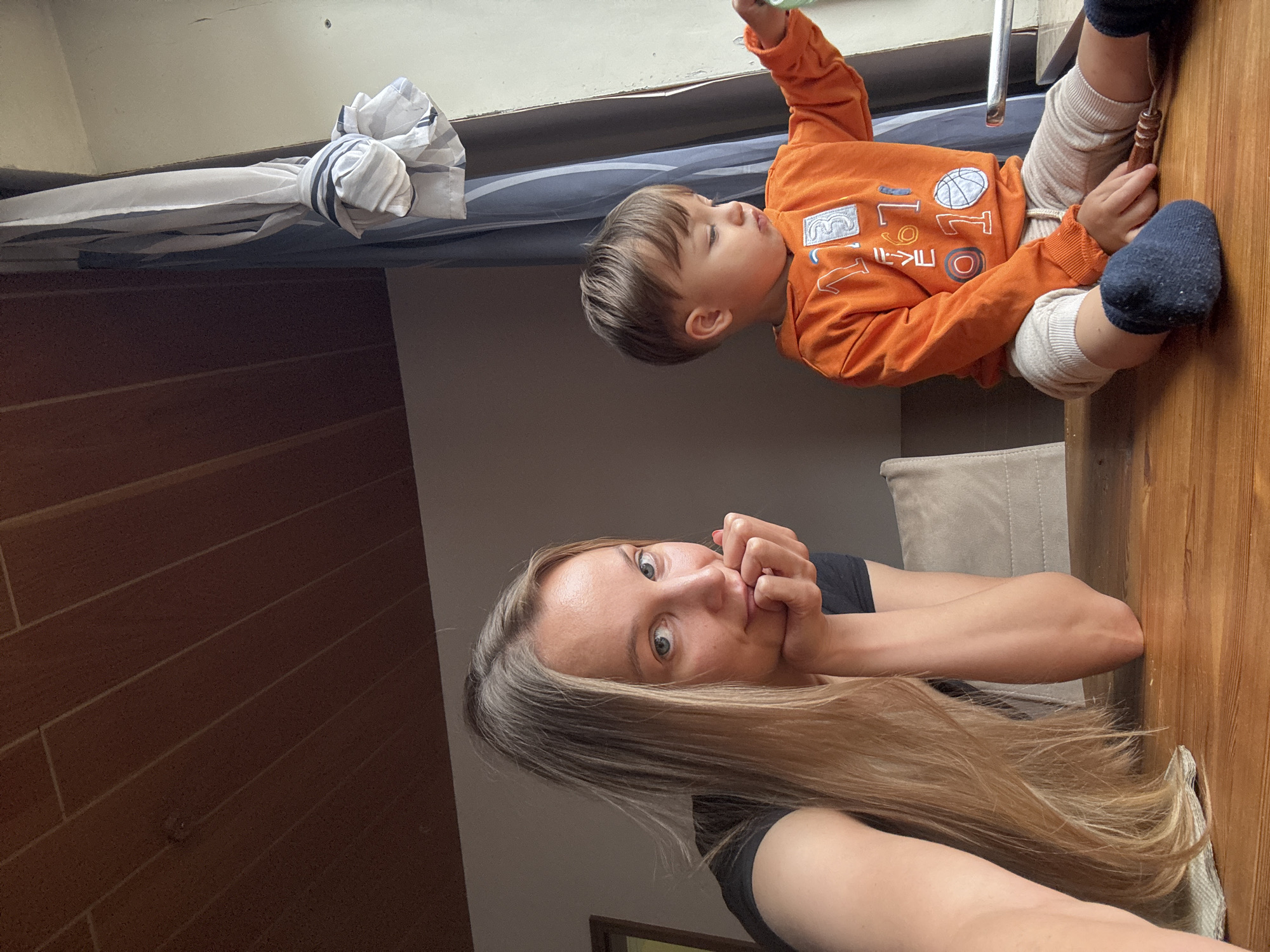 The image depicts a woman and a young child sitting at a wooden table. The woman, with long blonde hair, is in the foreground, resting her chin on her hand and looking towards the camera. The child, wearing an orange hoodie with graphics, is to the right, looking out of a window. The environment appears to be indoors, with a wooden paneled ceiling and a window with blue and white striped curtains. The scene is set within a residential interior. No specific location in Überherrn, Germany is discernible from the image.