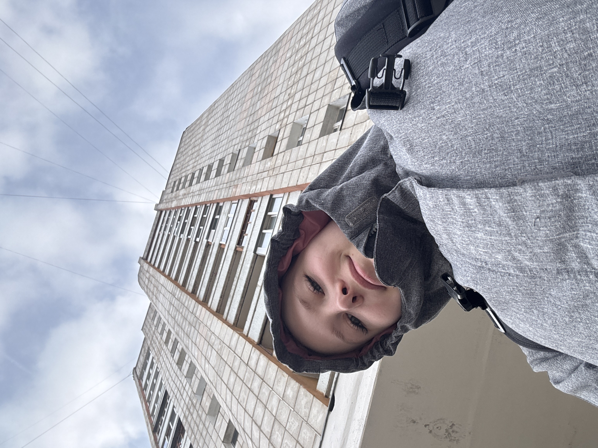 A low-angle, close-up photograph shows a person wearing a gray hooded sweatshirt. The hood is up, partially obscuring the person's face. Behind the person, a tall, gray-tiled building rises. Electrical wires stretch across a cloudy sky in the background. The building's facade features a grid-like pattern of windows and rectangular protrusions. A black buckle-type clasp is visible on the person's left shoulder. The location may be Chaikovsky, Russia, but there are no contextual clues to verify.