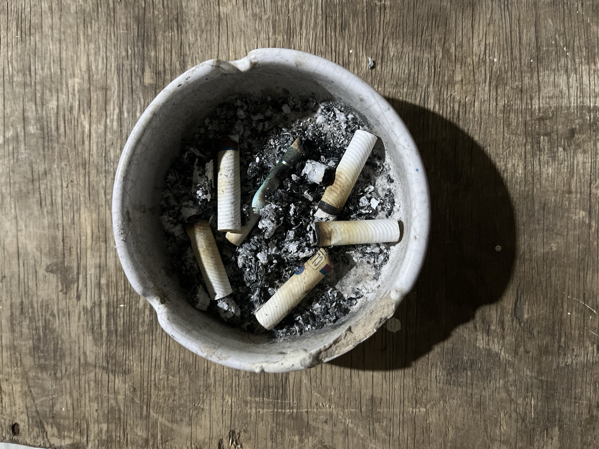 The image shows an ashtray filled with cigarette butts and ashes, placed on a weathered wooden surface. The ashtray is round and appears to be made of ceramic, with a chipped edge. Several cigarette butts are visible, some with burnt ends. The wooden surface has visible grain and a rough texture. The lighting suggests an indoor setting. There is no information in the image that provides context for the location in East Jakarta, Indonesia.