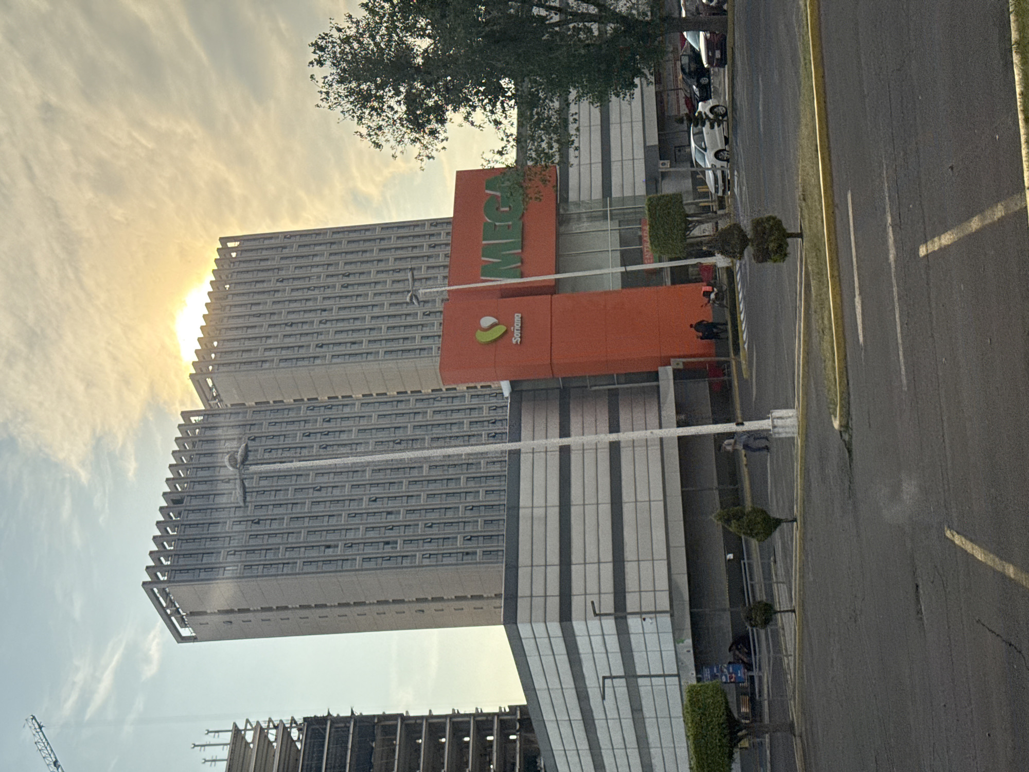 The image depicts a tall building complex in San Bernardino Tlaxcalancingo, Mexico, featuring a MEGA Soriana supermarket. The supermarket is recognizable by its orange facade and the large, green "MEGA" logo. Adjacent to the supermarket are two high-rise buildings, likely residential or office towers, with a uniform pattern of windows. In the foreground, there's a road with sparse landscaping. A construction site is visible at the bottom of the image, with cranes and incomplete building structures. The sky is bright with sunlight. There are a few people near the entrance of the supermarket. Cars are parked nearby.