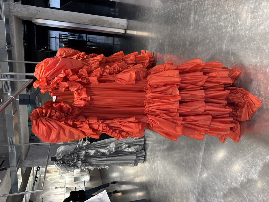 The image displays an interior view of a modern high-end fashion boutique or exhibition space in Paris, France, likely associated with Maison Margiela, suggested by a faint, reversed reflection of text on a dark surface in the background. The scene is dominated by a voluminous, coral-red dress, rich with multiple tiers of ruffles and gathered fabric, particularly prominent in its sleeves and skirt. This dress hangs from a black hanger on a polished metal rail in the foreground, extending nearly to the polished grey concrete floor, which reflects overhead lighting. To the left, slightly further back, a similar style dress in a metallic silver-grey color is also displayed on a hanger. In the background, a person is partially visible from the waist down, wearing dark pants and carrying a white bag, walking away from the camera. The environment features exposed concrete walls and pillars, contributing to an industrial-chic aesthetic. Overhead, metal rails and lighting fixtures are visible. The overall scene depicts a curated display of avant-garde fashion within a minimalist, contemporary setting.