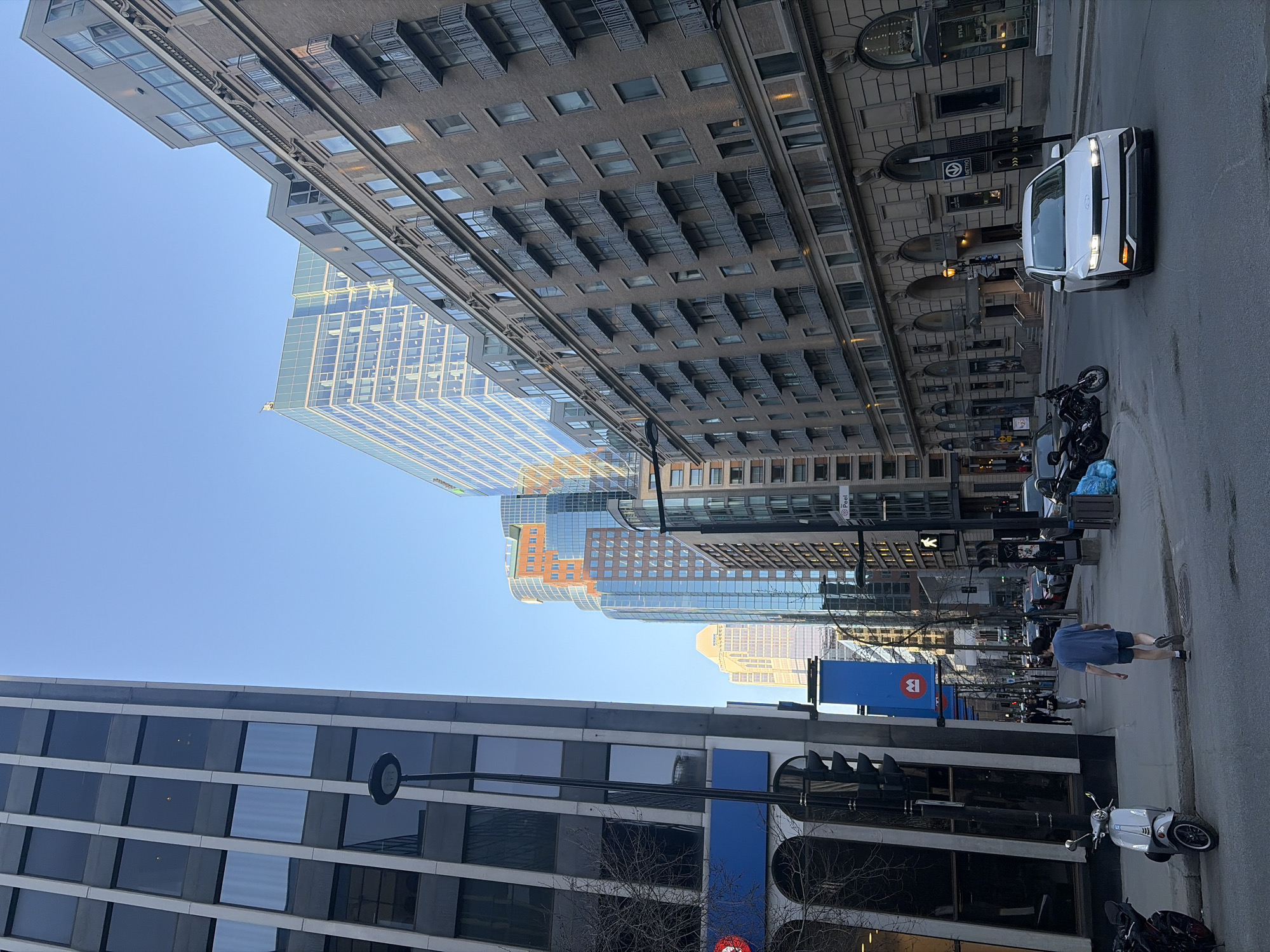 The image captures a vibrant street scene in Montreal, Canada, under a clear blue sky during the daytime. The street is lined with a blend of architectural styles. On the left and in the background, modern skyscrapers with reflective glass facades rise, showcasing their sleek designs. On the right, an older, more traditional building with an ornate stone or brick facade, archways, and detailed cornices stands prominently.
Vehicles include a white, modern SUV-like car (possibly a Hyundai Ioniq 5) on the right side of the street, with several motorcycles parked nearby. On the left, a white scooter, resembling a Vespa, is parked. Pedestrians are visible on the sidewalks; one man in a blue t-shirt and shorts walks on the right pavement.
Notable street furniture includes streetlights and a prominent blue pillar. Visible text on this pillar includes a red circle with a white "B", indicating the BIXI bike-sharing service. Additionally, a sign with a white "M" in a circle signifies a Montreal Metro station entrance. The bright sunlight creates sharp shadows and reflections on the glass buildings, highlighting the clear weather conditions. The scene suggests a typical day in a bustling urban environment.