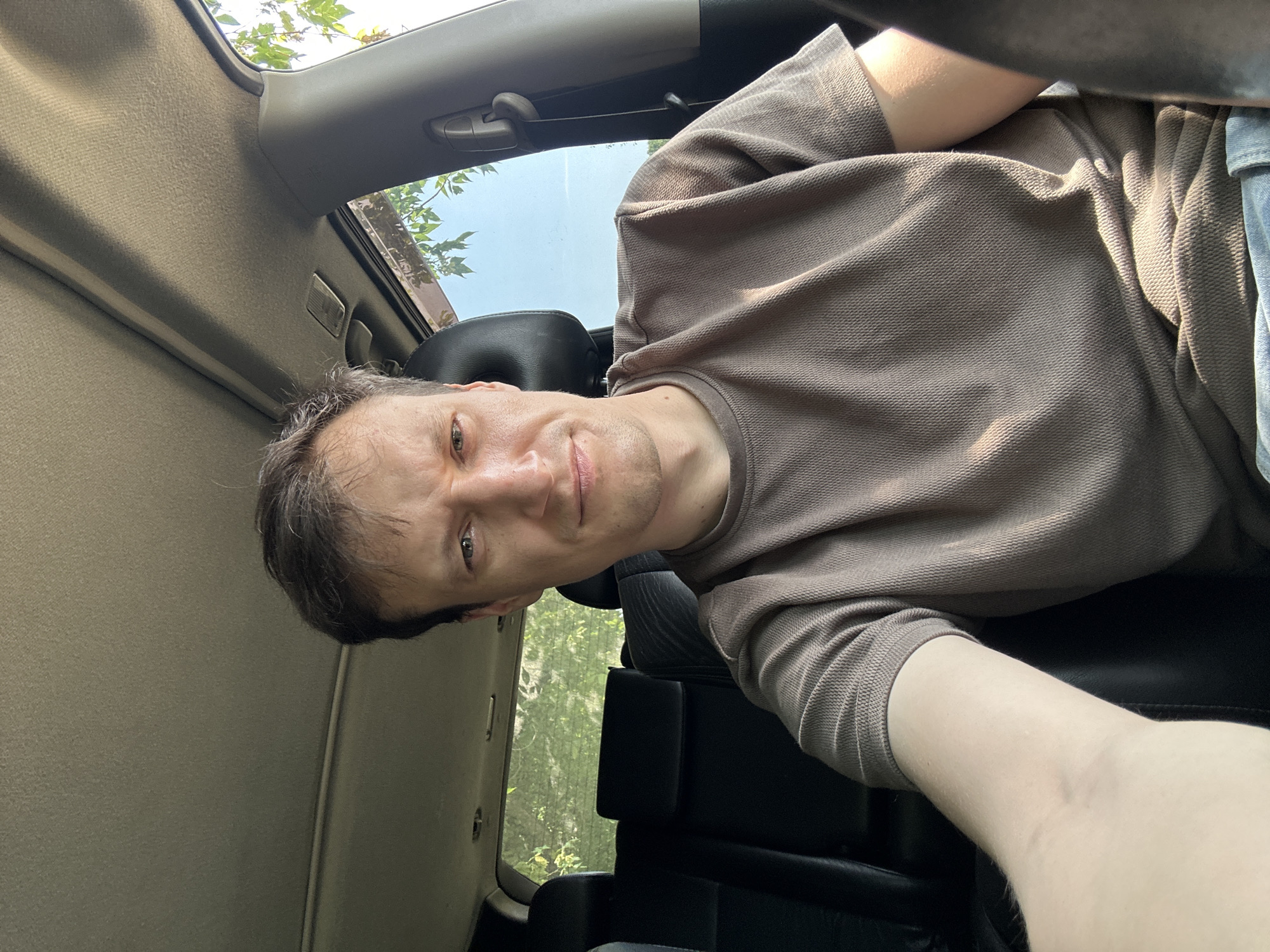 A man is sitting in the driver's seat of a car, facing the camera. He is wearing a brown t-shirt and blue jeans. The car's interior is visible, including the headliner, seats, and a window with trees outside. The man has short, dark hair and is looking directly at the camera with a neutral expression. The image appears to be a selfie taken in a sunlit environment. There are no visible signs indicating this is in the city of Makiv, Ukraine, or any other specific location.