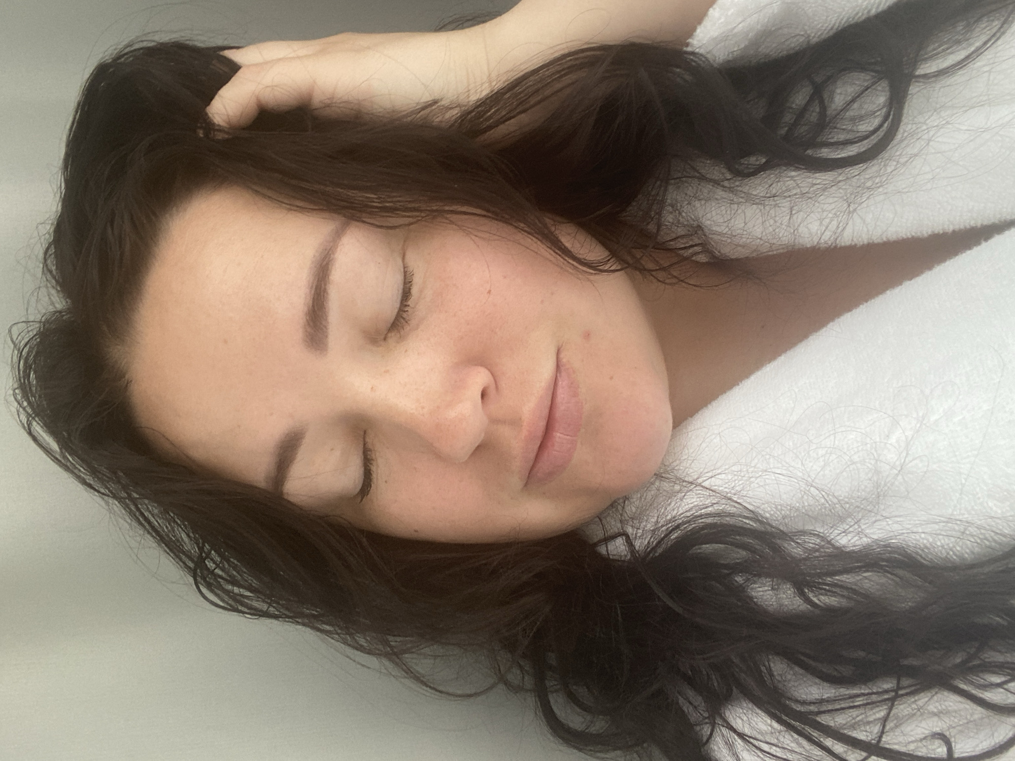 A woman with closed eyes and dark, wavy hair lies on a white surface, possibly a bed or spa table, wearing a white garment that could be a robe. Her hand is partially visible in her hair. The setting is indeterminate and there's no direct indication of being in Sochi, Russia.