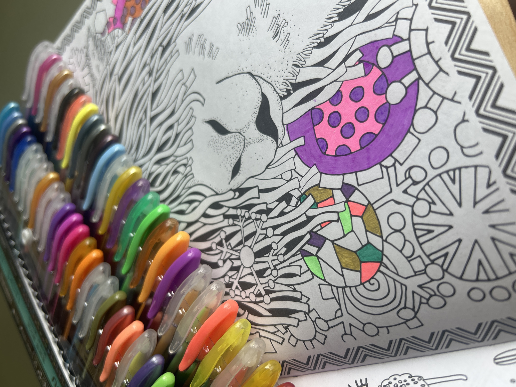 A close-up, high-angle shot captures an open adult coloring book alongside a collection of vibrant gel pens, suggesting an ongoing creative activity. The coloring book features intricate, abstract designs, with some sections partially colored in bright hues while others remain as black and white outlines.
On the right page of the book, a stylized design resembles part of a face, possibly a lion or a mythical creature, with wavy lines and stippled texture creating its features and mane. Adjacent to this, geometric and organic patterns are filled with color, including a prominent pink section adorned with purple polka dots, bordered by solid purple. Other completed areas showcase metallic gold, shades of green, and coral or light orange. A distinct snowflake-like pattern is also visible, currently uncolored.
To the left, a comprehensive array of gel pens is neatly organized in rows within a clear tray. The pens display a wide spectrum of colors, including various blues, greens, yellows, oranges, pinks, purples, and browns, with transparent caps revealing the vibrant ink. The pens appear ready for use, complementing the partially colored artwork. The scene suggests a hobby or leisure activity. The photo was taken in Izhevsk, Russia. No discernible text is visible in the image.