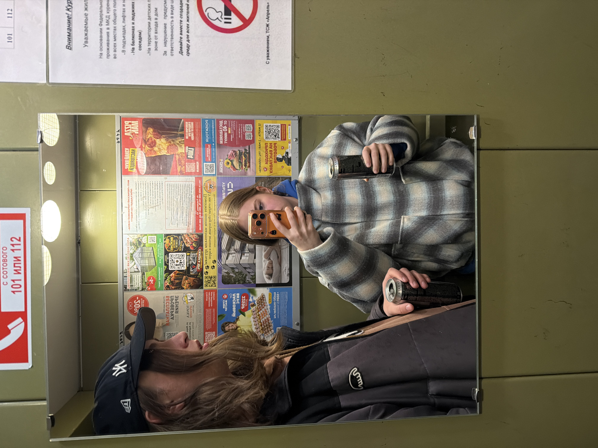 Two young people are reflected in a mirror that is likely in an elevator or public space in Perm, Russia, indicated by the Cyrillic script on the posters and signs surrounding the mirror. The person on the left is wearing a black New York Yankees baseball cap and a dark-colored jacket. The person on the right, presumably taking the photo with a cell phone, is wearing a blue shirt under a plaid coat and holding a black can. The mirror is covered with various advertisements and informational posters. The posters have bright colors and include images of food, QR codes, and Russian text. On the wall to the right of the mirror is a green surface, marked with a horizontal line. Above the mirror is a white poster with text in Russian, warning about smoking. A red and white sign with the numbers "101 или 112" (101 or 112) is also visible. The lighting is artificial and slightly diffused, giving a soft glow.
