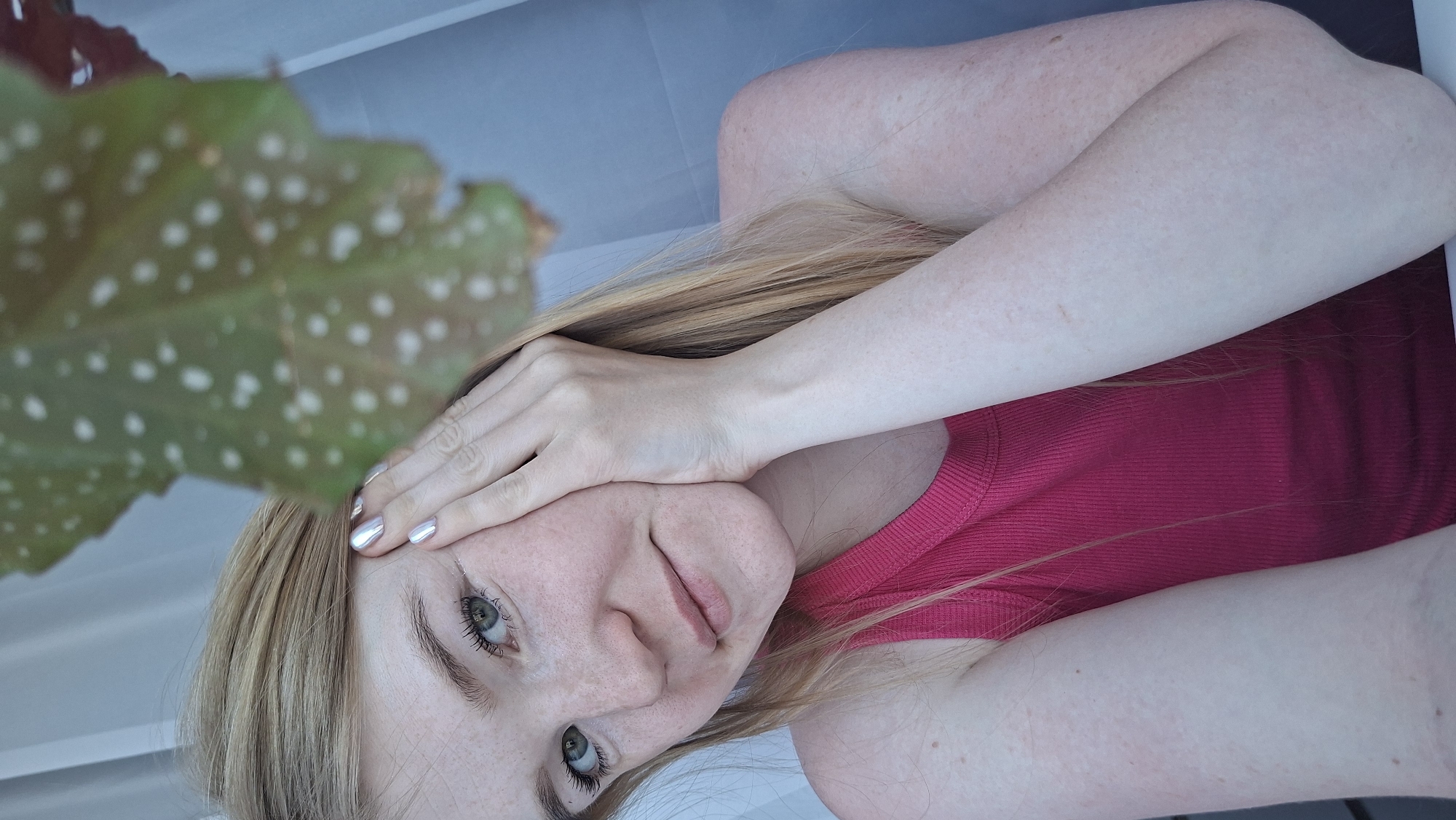 A young woman with blonde hair is facing the camera, with her head resting on her hand. She is wearing a red tank top. A large, green leaf with white spots is visible in the upper left corner of the frame. The background appears to be a light blue or gray color. The location is unknown and cannot be determined from the image.
