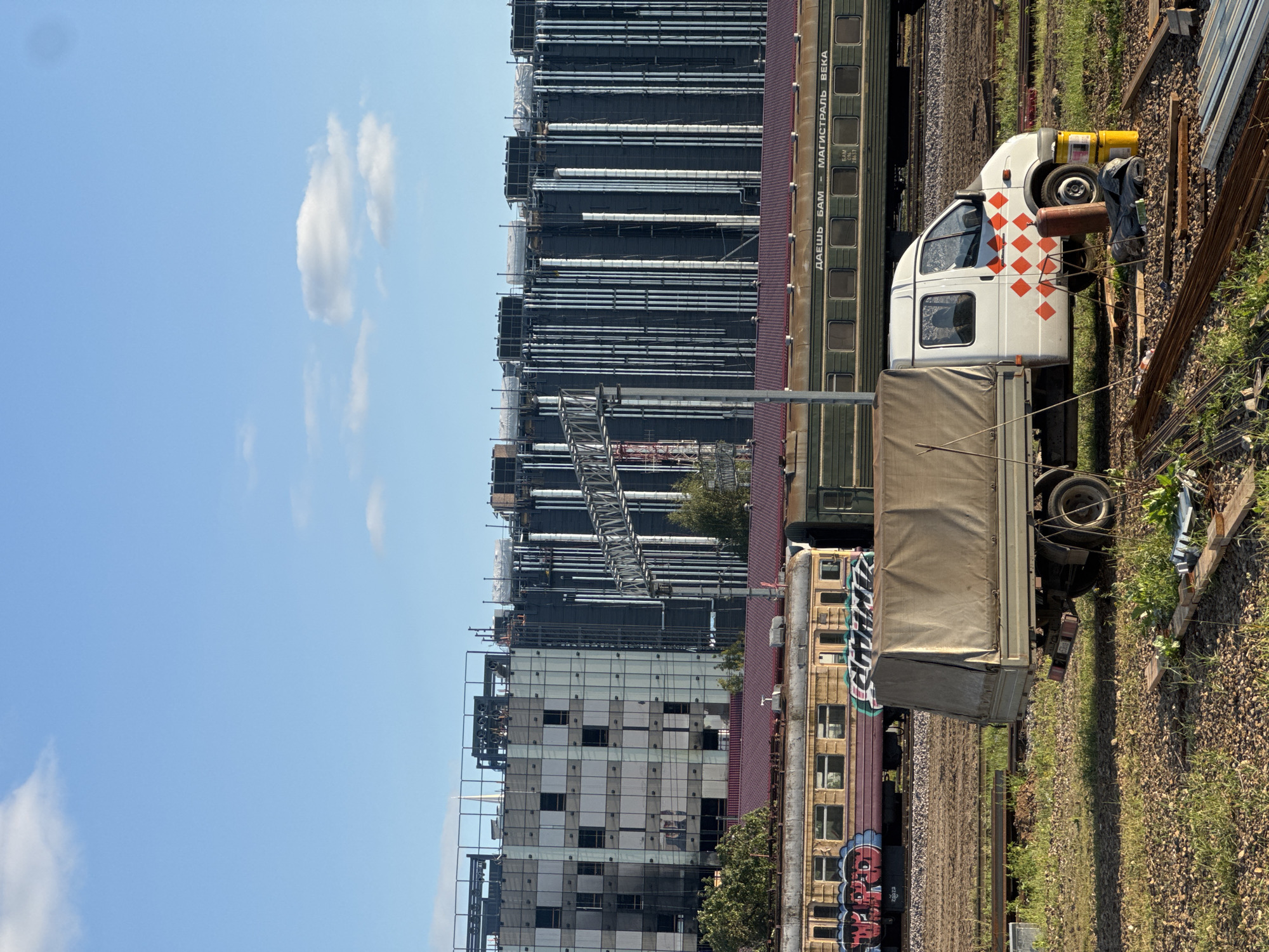 This is a medium-shot, outdoor, eye-level, vertical photograph taken on a sunny day in Khimki, Russia. In the foreground, a white flatbed truck with a beige canvas cover is parked on a gravelly, grassy area. The truck has red diamond patterns on its side. Next to the truck are a red propane tank, a yellow barrel, and scattered construction materials, including rebar and wooden planks.
In the middle ground, two train cars are parked on parallel tracks. The closest train car is yellow with red and blue graffiti on its side. The second train car is green and has Russian text on its side which reads "ДАЕШЬ БАМ – МАГИСТРАЛЬ ВЕКА".
In the background, a large, modern building under construction is visible. The building has a dark facade with rows of vertical silver pipes and numerous square windows. The sky above is bright blue with a few scattered white clouds.