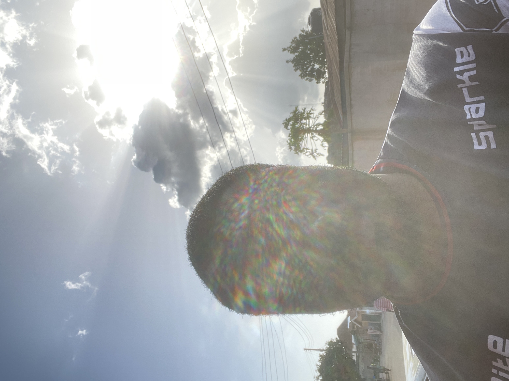 The image shows a close-up view of a person's head and upper body, partially obscuring the sky. The person has short, dark hair. They are wearing a black shirt with the word "Sparkle" printed vertically in white on the right side. The background features a bright, sunlit sky with clouds and sun rays. There is a building to the right and trees and buildings are visible in the distance. Considering the information and the likely source of images, the location is consistent with Jos, Nigeria.