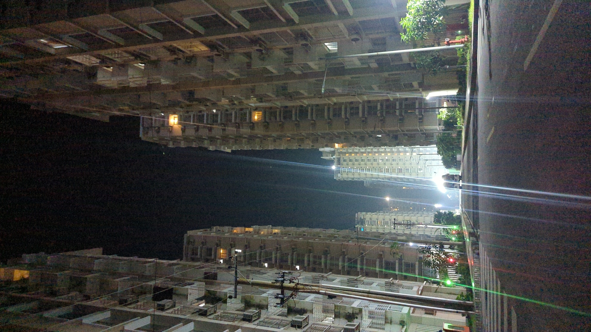 The image depicts a nighttime street view in Chor Sahapur, Bangladesh, characterized by the presence of numerous tall apartment buildings. The buildings, illuminated from within by lights in their windows, flank a paved road. In the foreground, a crosswalk is visible. A vehicle is present on the road in the distance, with its headlights on, contributing to the bright streaks of light across the scene. Streetlights and utility poles with wires are also visible. The overall impression is one of an urban residential area at night, with the architecture dominated by multi-story apartment complexes.