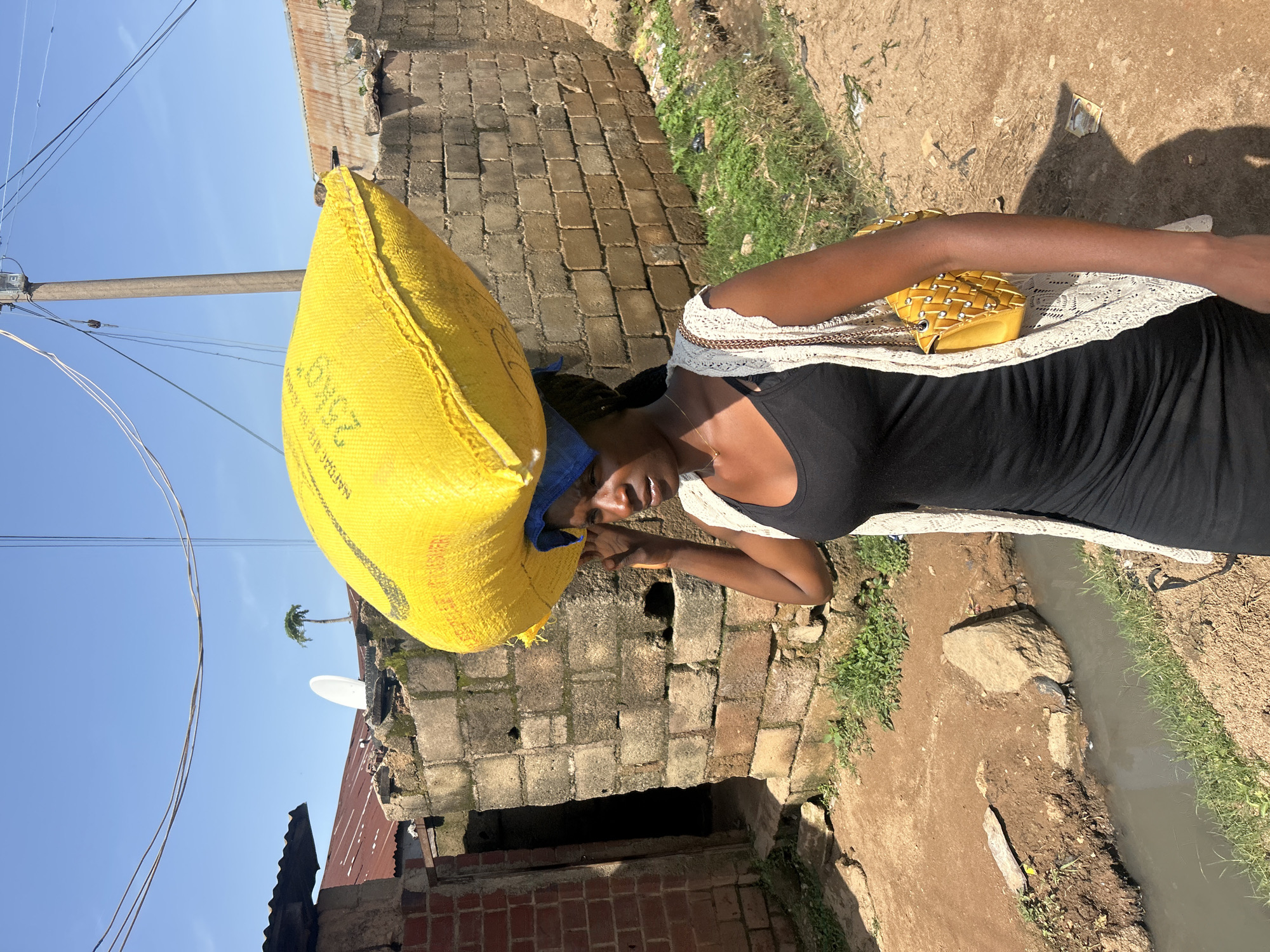 A medium shot captures a woman walking outdoors in Jos, Nigeria, with a large, bright yellow sack balanced on her head. She is wearing a black sleeveless dress and a cream-colored, open-weave cardigan. A yellow purse with a quilted pattern and studded embellishments is slung over her right shoulder. She has dark hair and is looking slightly down and to her right, with her right hand adjusting the sack on her head.
The environment appears to be a dusty, unpaved area with some vegetation and a small, murky stream or drainage ditch running alongside the path. In the background, there are brick structures with corrugated metal roofs, characteristic of urban or peri-urban settlements in Jos. A satellite dish is visible on one of the rooftops, and power lines stretch across the bright blue sky. The overall impression is of a scene in a residential or market area within the city.