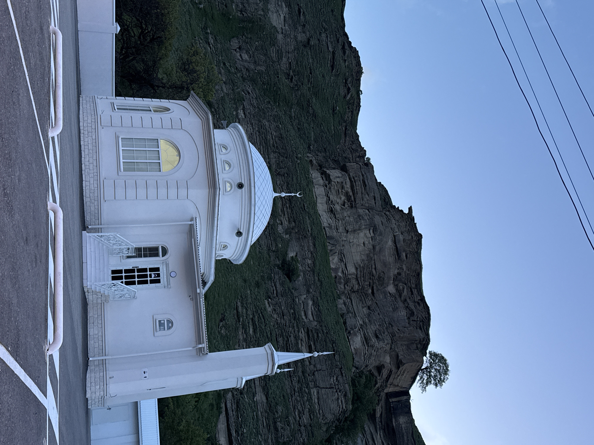 The image shows the white mosque located in the city of Ordzhonikidzevsky, Russia. The mosque stands in front of a large, rocky mountain covered with vegetation. The structure features a white dome topped with a crescent moon symbol, and a minaret. Power lines are visible against the blue sky. The mosque is adjacent to a paved road marked with white lines.