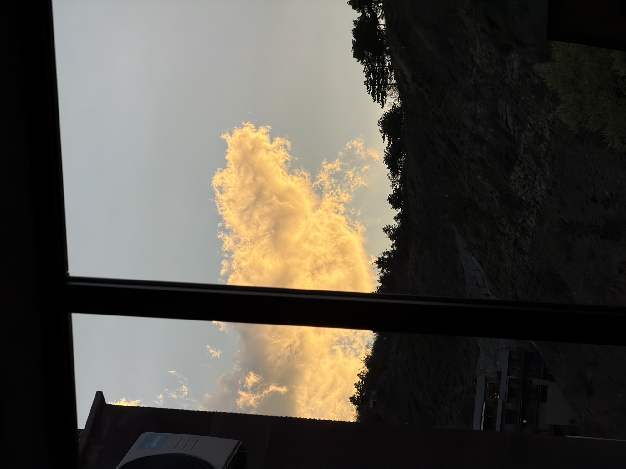 The image shows a view from inside a building in Tbilisi, Georgia, looking out a window at a bright cloud lit by golden light against a pale sky. A steep, dark rock face covered with trees is visible to the right of the cloud. Part of another building is visible at the bottom of the rock face. The interior is dimly lit, with a black window frame in the foreground. An air conditioner unit is visible at the bottom left. The time of day appears to be either sunrise or sunset.