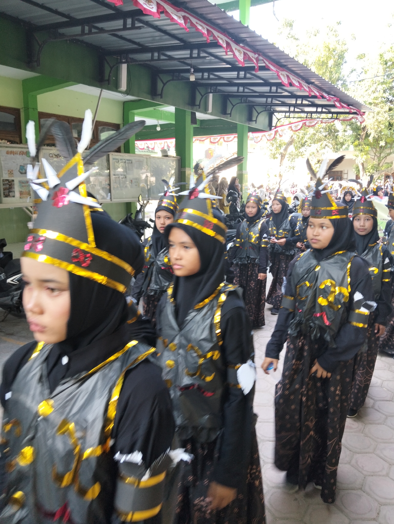 In Kranji, Indonesia, a group of young girls in traditional attire with feathered headpieces and black and gold vests are walking in a procession. They are wearing black hijabs and patterned black skirts. The procession appears to be part of an outdoor event, possibly a parade or festival, as evidenced by the banners overhead and the tiled pathway. The environment includes a building with green walls and a covered walkway. Several motorcycles are parked in the background. The lighting suggests it is daytime with bright sunlight.