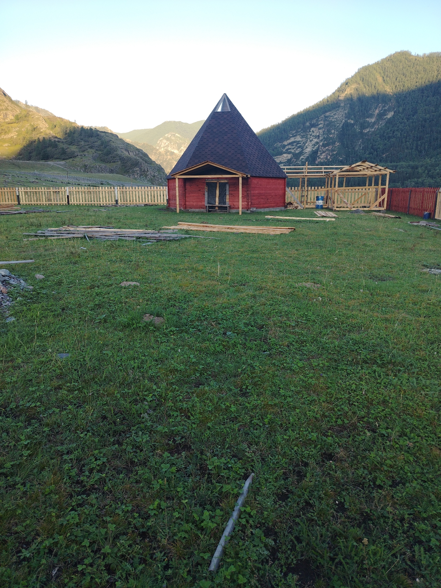 In Iodro, Russia, a small red wooden structure with a conical roof sits on a grassy area. A light-colored wooden fence surrounds the property. In front of the structure, there is a partially constructed wooden gazebo. Lumber is piled on the grass, suggesting ongoing construction. The background features a steep, forested mountain slope under a clear sky.