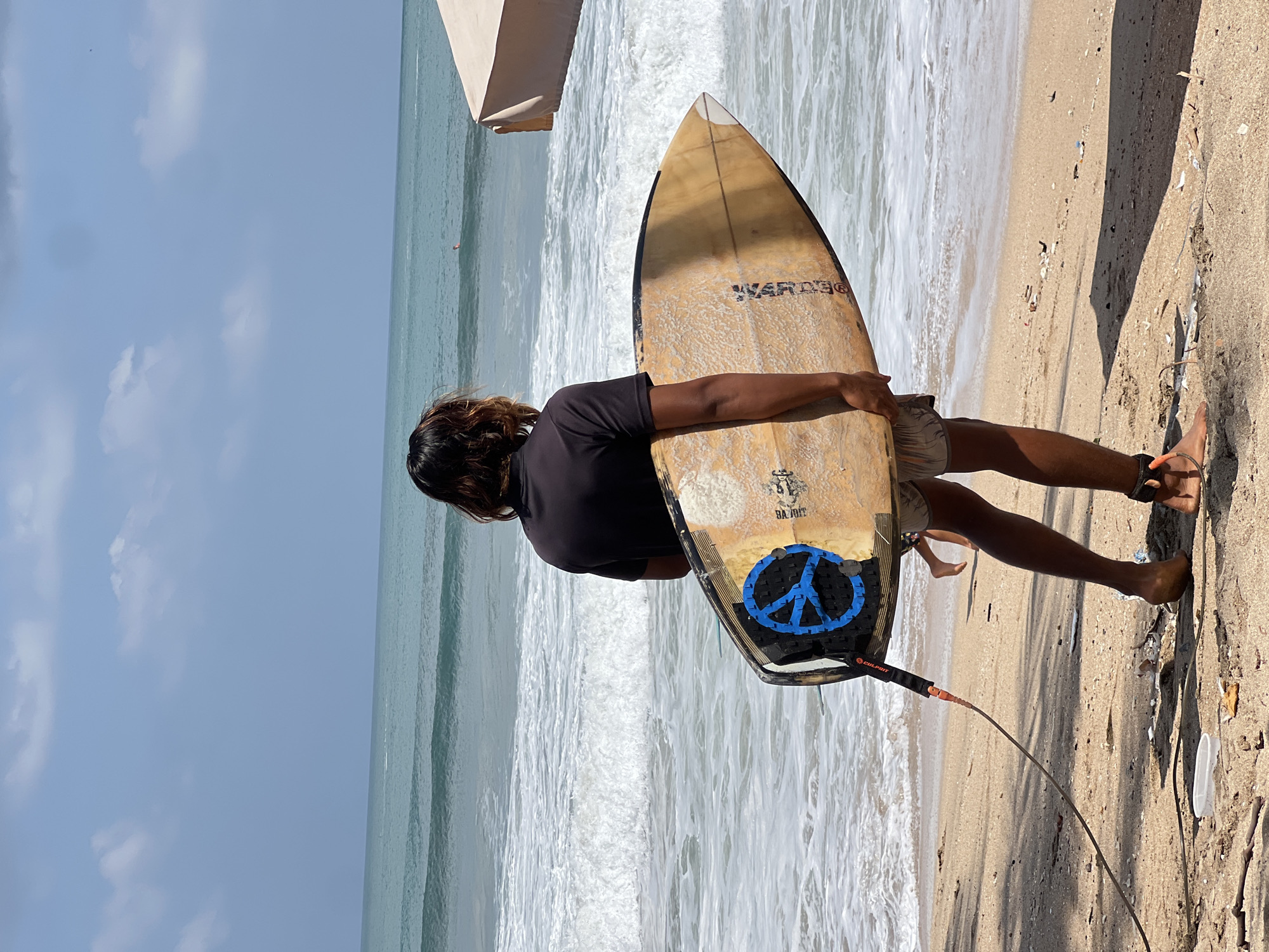 In Kuta, Indonesia, on a sandy beach, a surfer stands with their back to the camera, holding a surfboard. The surfer wears a black t-shirt and light-colored shorts, with a leash attached to their ankle and the surfboard. The surfboard itself is light brown with a blue peace sign decal on its deck. The ocean, with moderate waves and white foam, stretches out before them under a partly cloudy sky. In the distance, a person can be seen in the water. To the right, a portion of a light-colored beach umbrella is visible. The beach is littered with small debris.