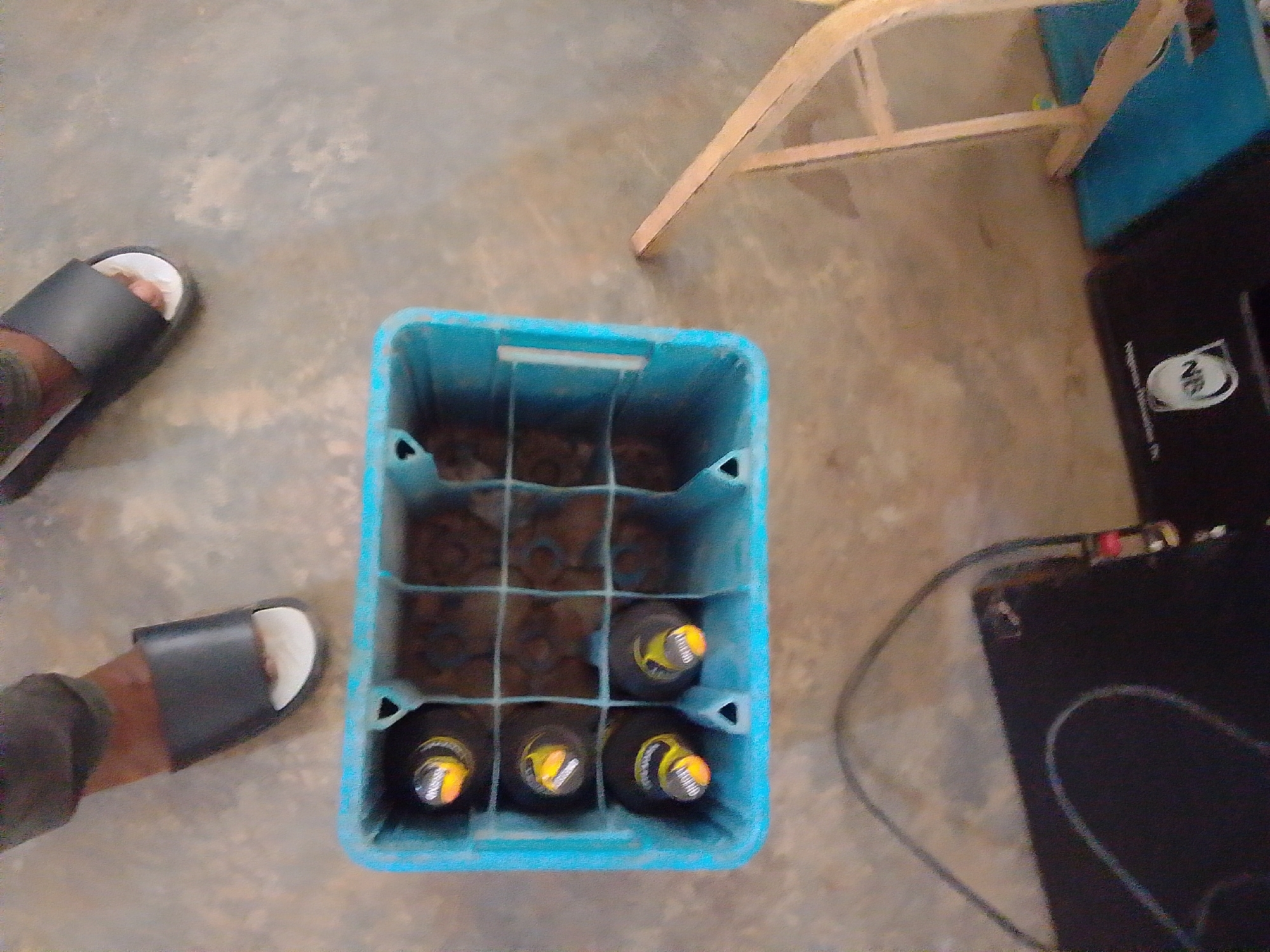In New Stadium, Nigeria, a ground-level, slightly high-angle shot captures a scene on a concrete floor. In the foreground, a blue plastic crate is filled with several dark bottles, their yellow and orange caps visible. To the left, a person's feet are visible wearing black flip-flops, one on the left foot with a white toe visible, and another on the right foot. The person is wearing dark pants. In the background, a wooden stool-like structure with lighter wooden legs is positioned over a dark object, possibly electronic equipment, with a black cable snaking away. The lighting suggests an indoor or shaded outdoor environment.