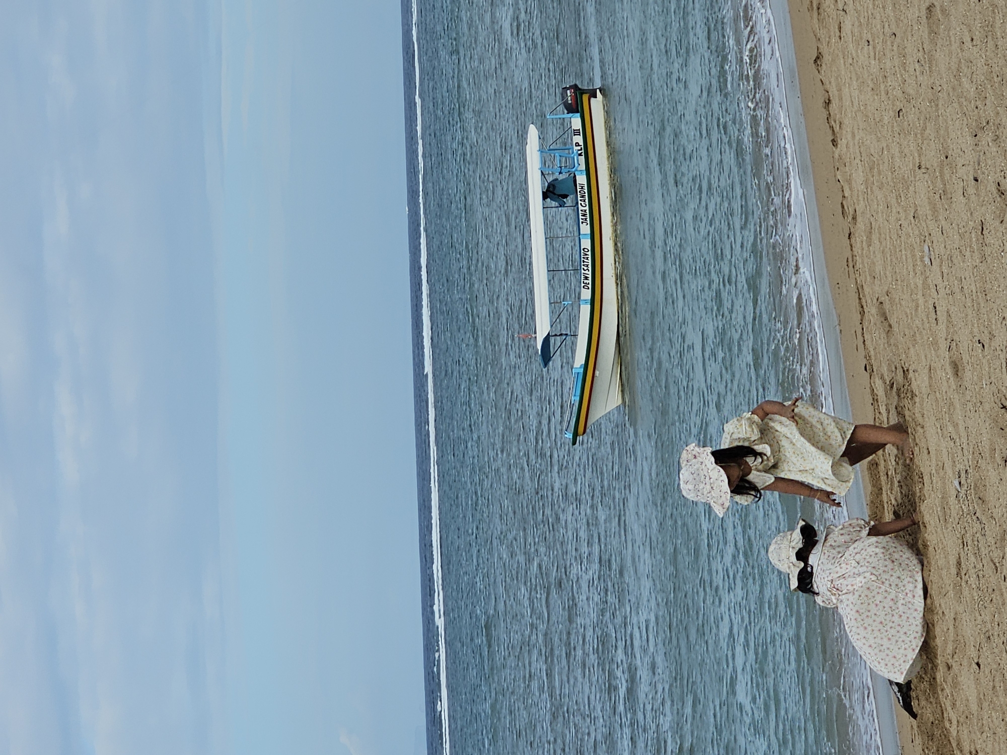 The image depicts a beach scene with two young girls in matching floral dresses and sun hats playing on the sand near the water's edge. In the background, a white boat with a blue canopy is on the sea, with the text "DEWI SATAYO JANA GANDHI KLP III" visible on its side. The water is a deep blue with whitecaps, and the sky is a pale blue with scattered clouds. The sand is light brown and shows subtle footprints. This scene is consistent with beaches found in Denpasar, Indonesia, a coastal city on the island of Bali known for its beautiful shores and maritime activities.