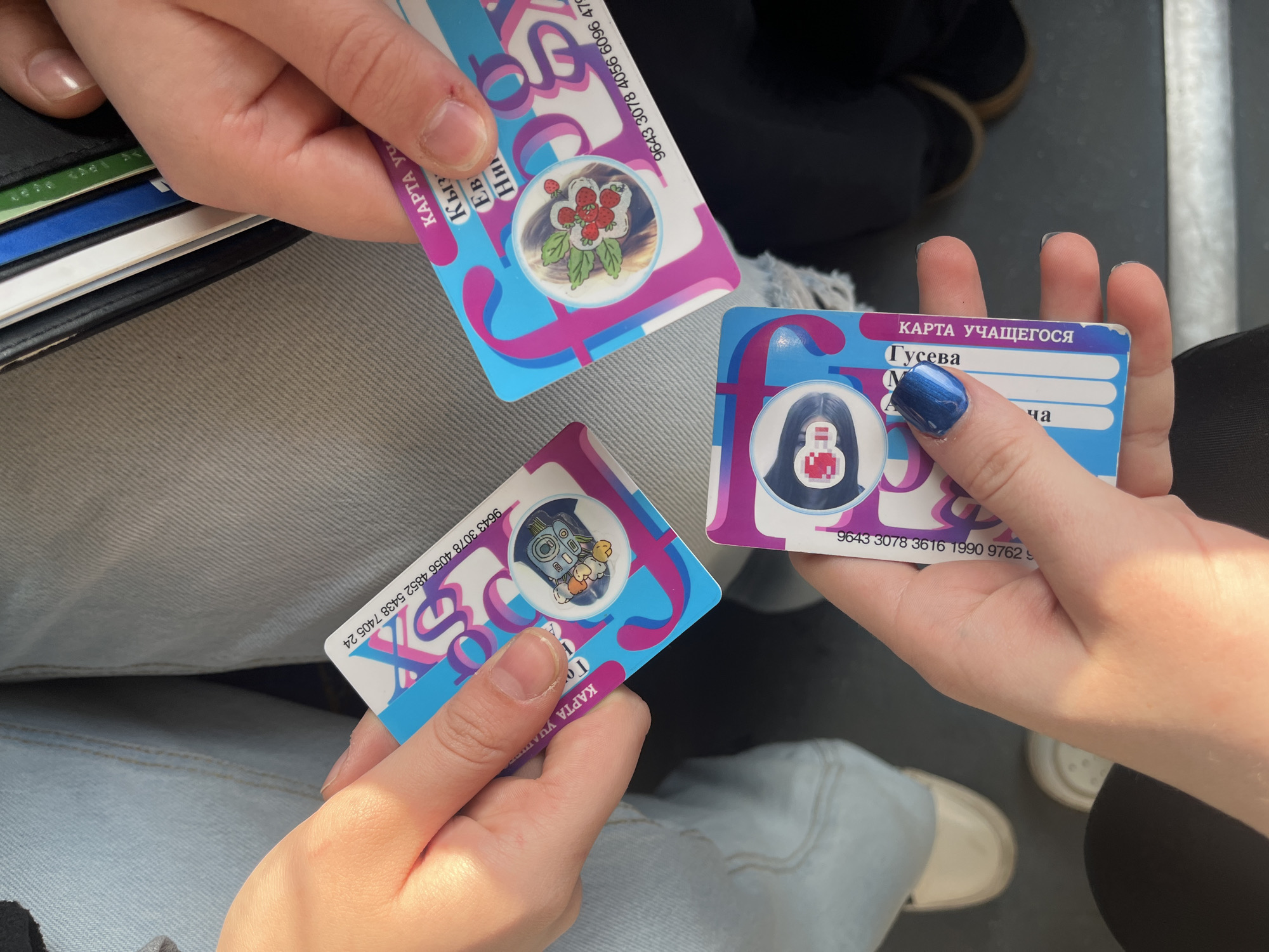 Here is a detailed description of the image:
**Overall Scene:** The image shows a close-up, slightly overhead view of three hands holding student cards. The background suggests they might be inside a public transportation vehicle, possibly a bus or tram, based on the visible floor and seating.
**People:** Three individuals are present, but only their hands and lower portions of their clothing are visible. One person has dark blue nail polish on their nails.
**Subjects/Objects:**
* **Student Cards:** Each hand holds a "Karta Uchashchegosya" which translates to "Student Card" in English. The cards have a white base with blue and purple stylized letter "f" designs. Each card has a different sticker/decoration on the photo area.
* **Personal Items:** One hand is near a black wallet with various cards inserted, suggesting payment cards or ID.
**Actions/Interactions:** The main action is the holding and displaying of the student cards. The three individuals appear to be interacting, possibly comparing or showing off their cards.
**Environment:** The setting appears to be inside a public transportation vehicle. The floor is a dark color, and the seats are upholstered in light-colored material.
**Additional Details:**
* The cards have serial numbers visible along the top edge.
**Location:** Based on the design and Cyrillic text on the cards, the location is most likely in Russia, specifically Saint Petersburg. The "Karta Uchashchegosya" is a specific type of card used for student transportation in that city.