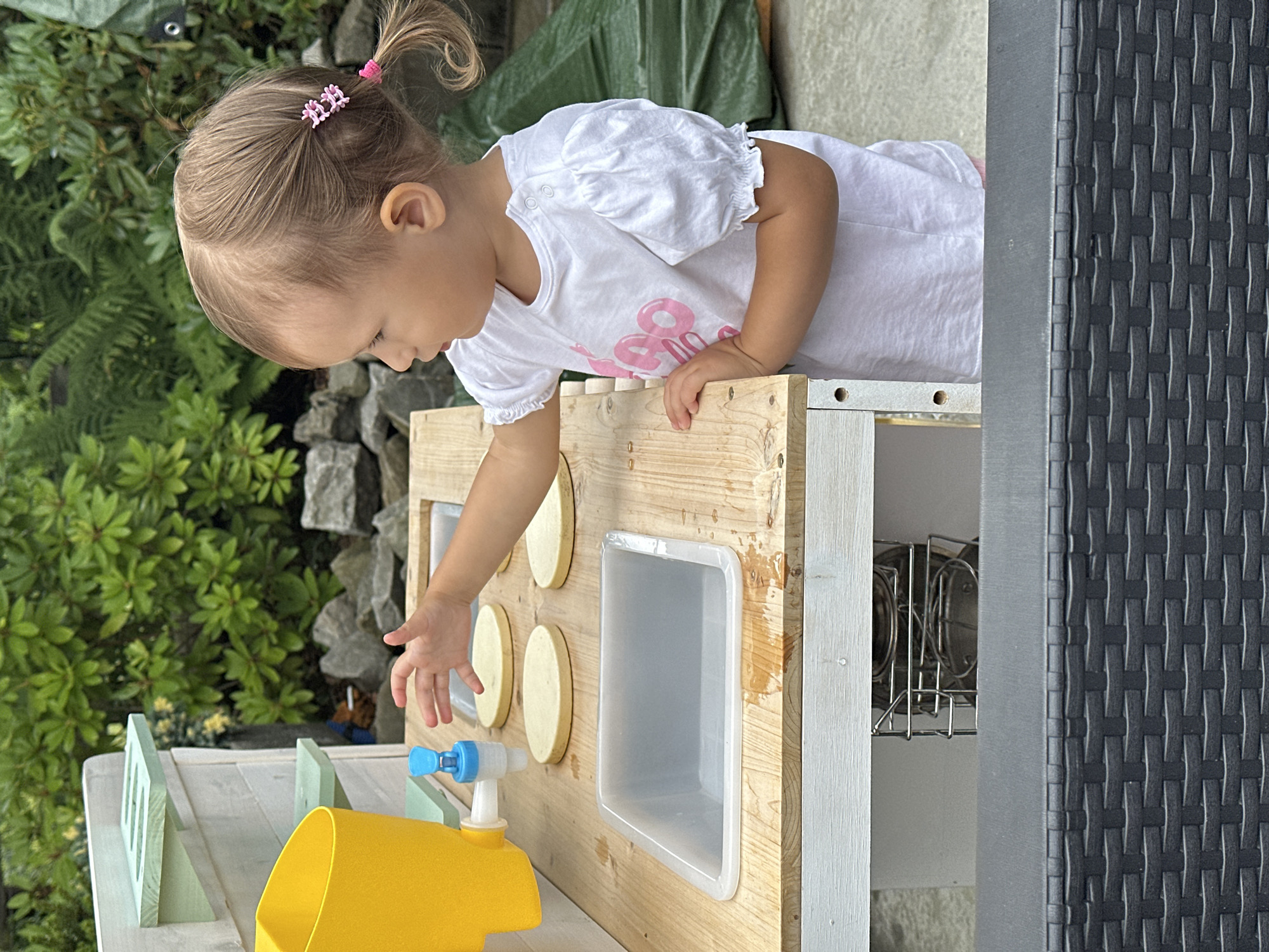 An outdoor scene captures a young girl of Caucasian ethnicity, appearing to be between two and three years old, engaged with a toy kitchen. She has fair skin, light brown hair pulled into a ponytail with pink accessories, and is wearing a white t-shirt with pink lettering. Her left hand is extended towards a blue faucet on a yellow water dispenser, suggesting she is interacting with it. The toy kitchen is made of wood and painted white and light green, featuring shelves, a sink, and circular wooden pieces that resemble plates. The background consists of lush green foliage, rocks, and a green tarp, indicating a garden or outdoor recreational area. The foreground shows a portion of a dark-colored woven outdoor furniture piece. The image does not provide specific information about the city of Wisła, Poland.