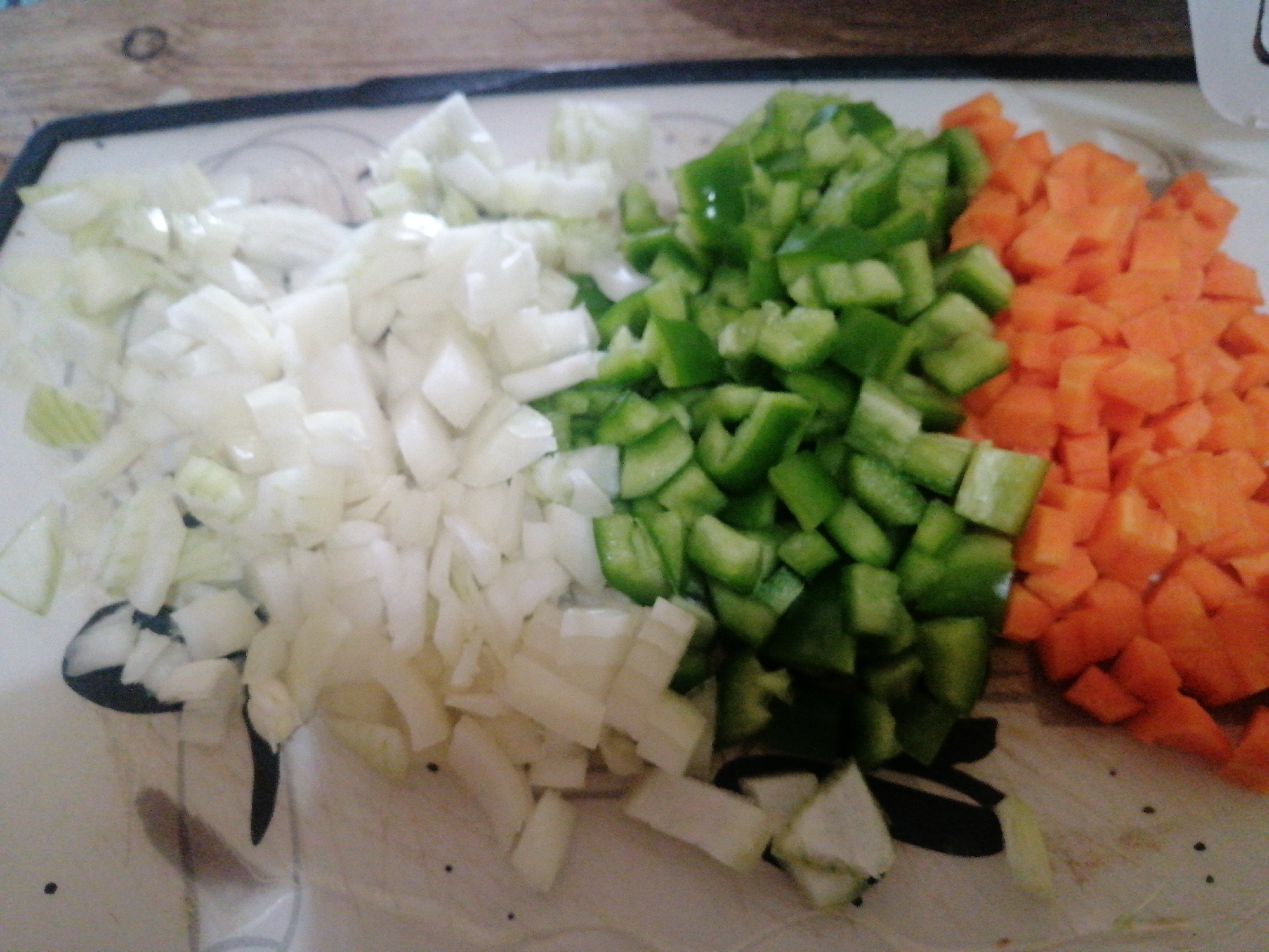The image shows a close-up of diced vegetables on a cutting board. From left to right, there's a pile of chopped white onions, a pile of chopped green bell peppers, and a pile of chopped orange carrots. The cutting board appears to be made of light-colored plastic. The vegetables are all roughly the same size. The background is blurred, with a wooden surface visible. The location is impossible to determine from this image.