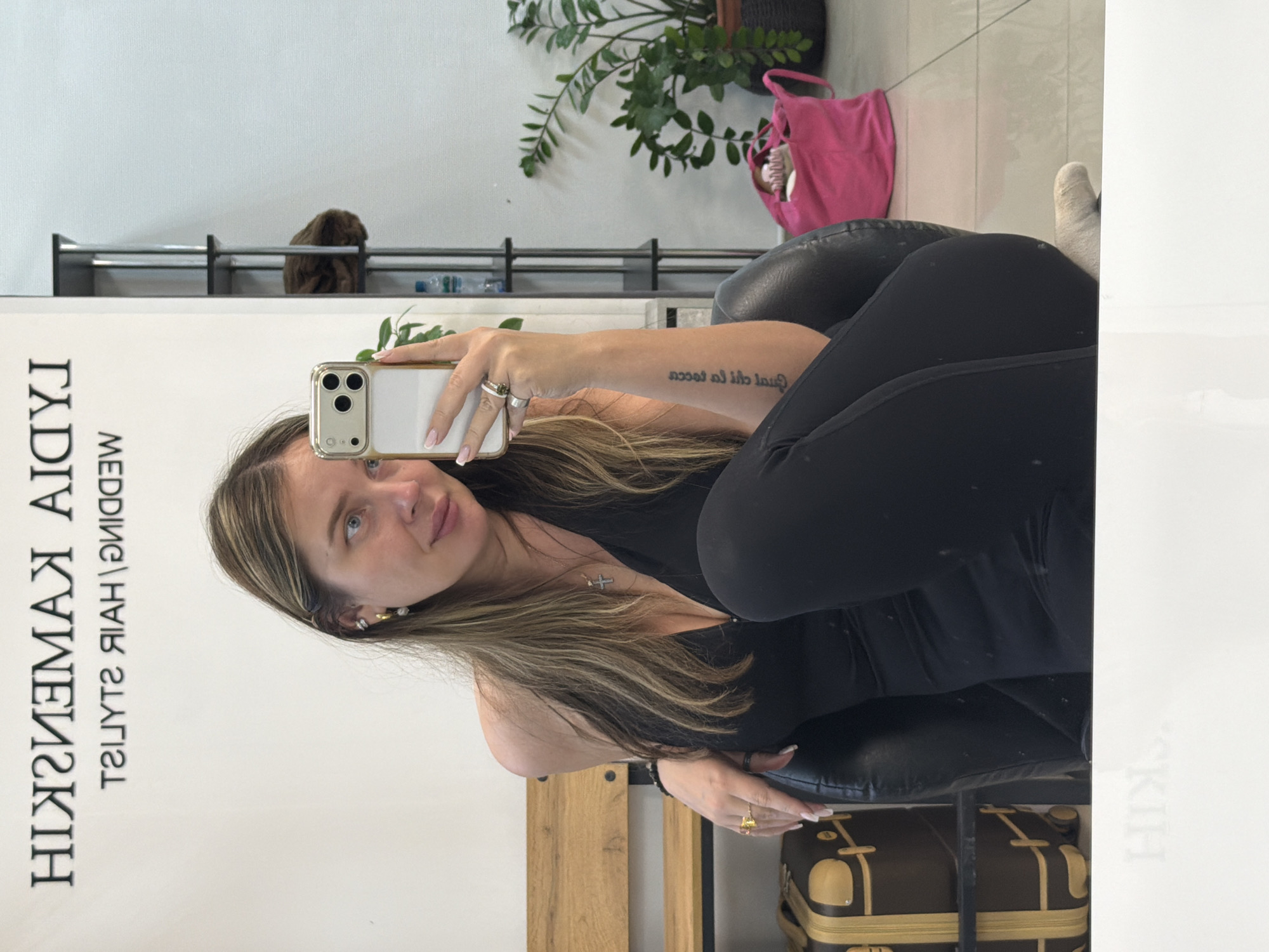 A woman is taking a selfie in a room with a large mirror. The mirror reflects the text "HIKIZMEMAX AIDA" in reverse, with the additional text "WEDDING\HAIR STYLIST" underneath. The woman is holding a smartphone and partially obscures the text in the mirror. She has blonde hair with darker roots, and is wearing a black top and dark leggings. She is seated on a black leather chair. The environment includes a plant, a pink bag, a shelf, and luggage-like storage containers. The location may be in Chaikovsky, Russia, but it cannot be conclusively determined from the image alone.