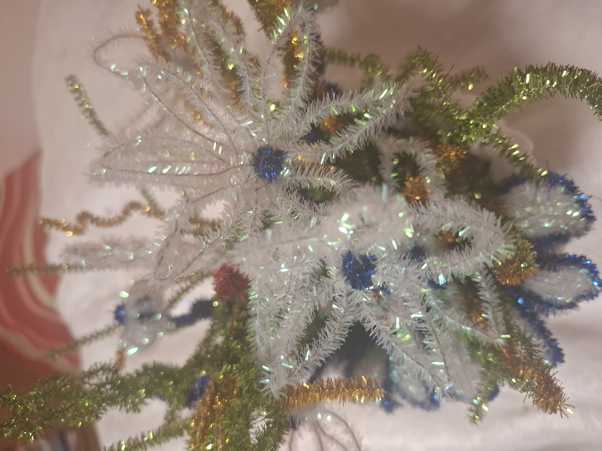 The image displays a close-up, slightly out-of-focus view of a festive arrangement, possibly a small Christmas tree or a decorative centerpiece. The arrangement is composed of various metallic pipe cleaners and tinsel in colors including white, green, gold, and blue. Some sections are shaped like stars or snowflakes, while others resemble evergreen branches. Small, spherical blue ornaments are interspersed throughout the arrangement, along with a few red accents. The background is soft and out of focus, suggesting a fabric surface, possibly a tablecloth or blanket, with hints of a striped pattern in the upper portion of the image. There is no visible indication of the city of Mıñbaev, Kazakhstan, or any specific environmental details that would place the scene there. The lighting appears to be artificial, casting soft highlights on the metallic elements. The overall impression is one of holiday decoration.
