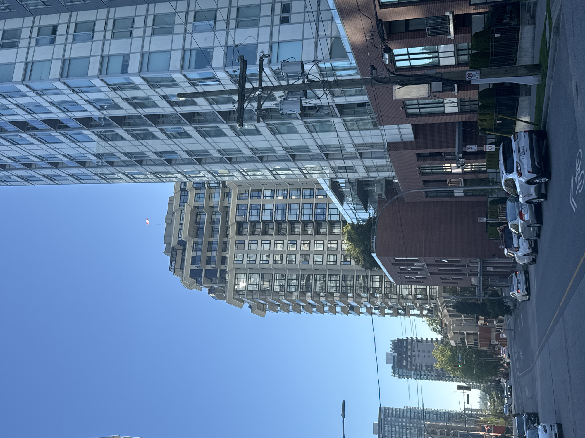 This is a street-level, medium-distance, eye-level, full shot of a city street in New Westminster, Canada, on a clear day. The perspective is looking down a street lined with modern, multi-story residential buildings.
On the right side of the frame, a tall, modern apartment building with a glass facade dominates the scene. Adjacent to it, closer to the foreground, is a brick building with large windows and balconies. A wooden utility pole with transformers and wires stands prominently in front of these buildings.
On the left side of the street, more tall apartment buildings are visible in the distance. The street itself is paved and has lane markings. Several cars are parked along the side of the road and driving in the distance.
In the sky, above the buildings, a Canadian flag is visible, small and red and white, atop one of the taller buildings in the middle ground. The overall impression is of a contemporary urban environment.