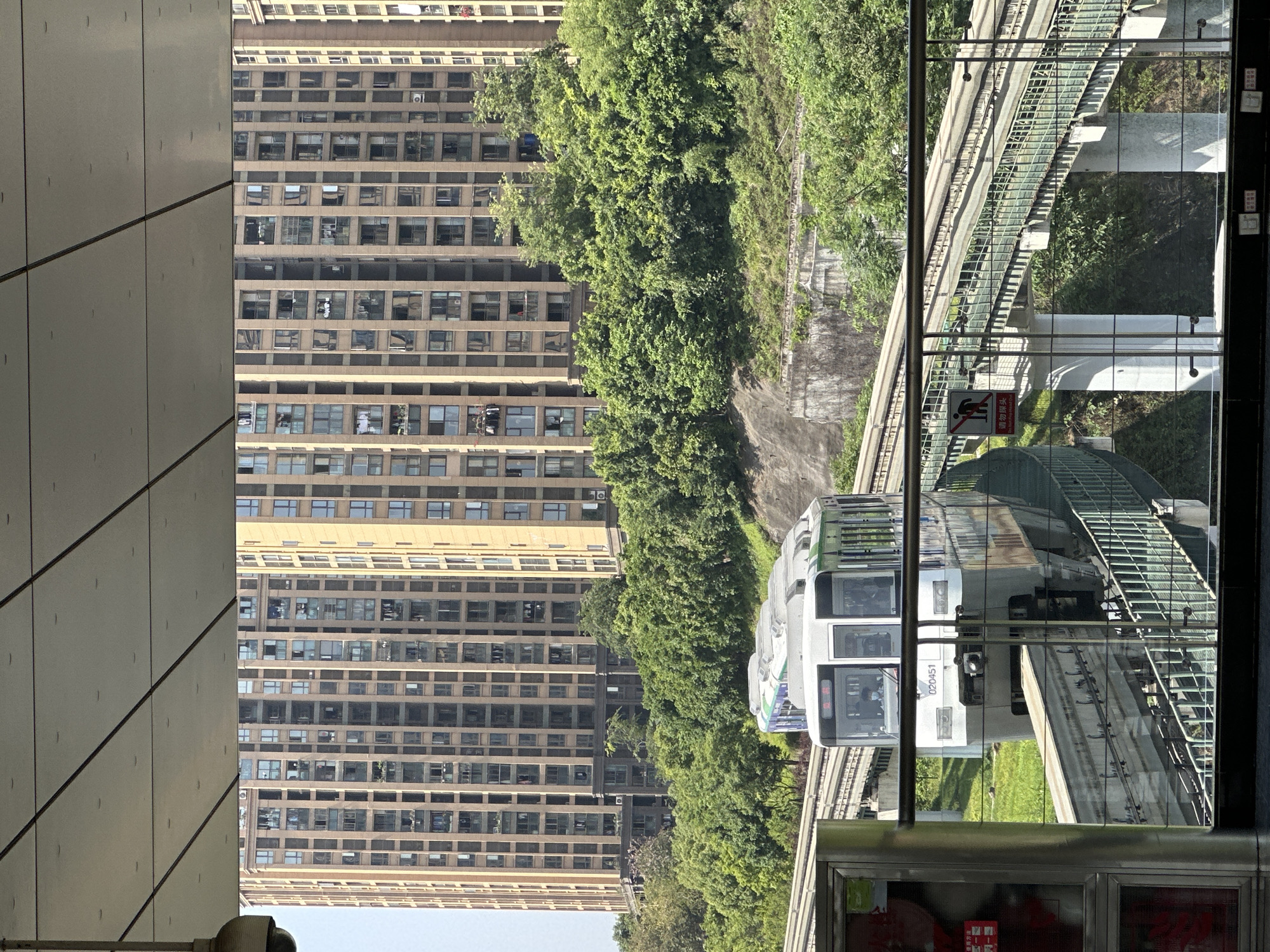 A monorail train is traveling along an elevated track in Jiansheng, China. Dense green trees line the track. A tall building with multiple windows can be seen to the left of the trees. The image is taken from inside a building, as part of the structure frames the view. The train is white with gray and green accents. The area around the tracks appears to be grassy.
