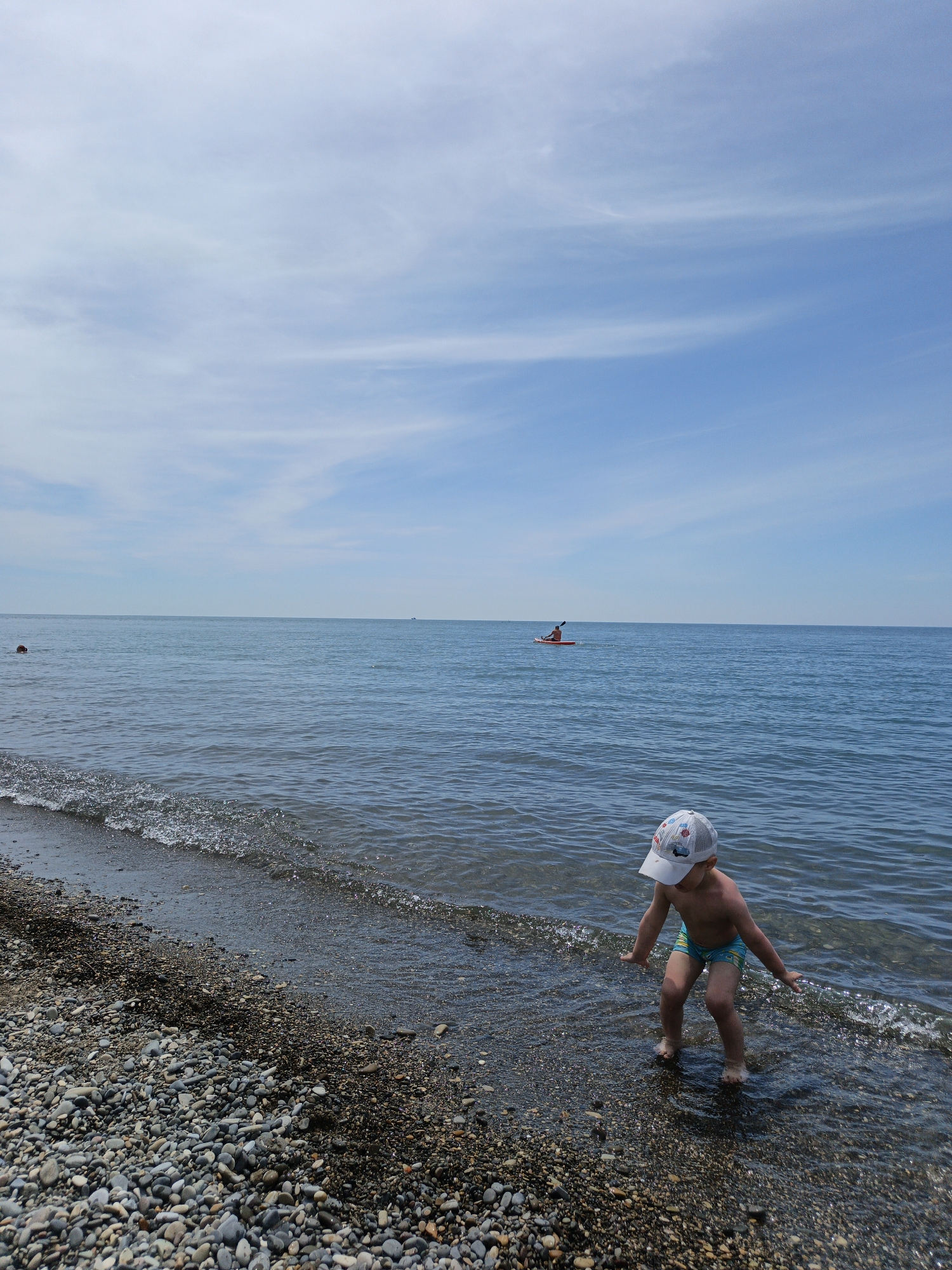 This vertical photograph captures a daytime scene at a pebble beach in Krinitsa, Russia. In the foreground, a young child wearing light blue swim trunks and a white baseball cap with colorful designs stands in the very shallow water at the edge of the dark, multi-colored pebble beach. The child is bent forward with arms slightly spread, facing the water, with small waves breaking around their bare feet. In the middle ground, a person in an orange kayak paddles leftward across the calm, blue-grey sea, creating a subtle wake. Further to the left, a swimmer's head and shoulders are partially visible above the water. The upper portion of the image displays a clear blue sky with sparse, wispy white clouds. The horizon line is flat, separating the calm sea from the sky.