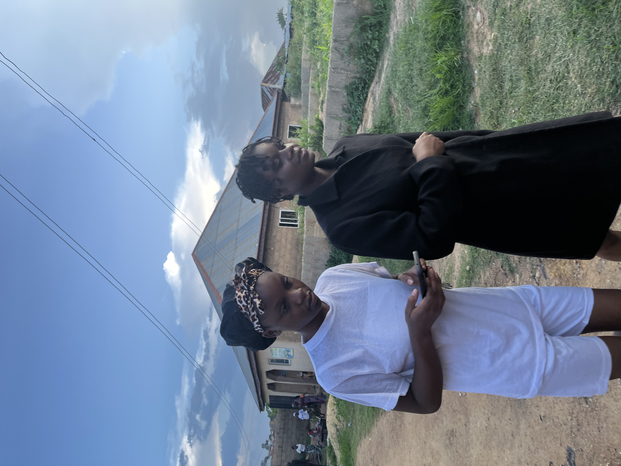 Two young Black women are walking outdoors in a village setting. The woman on the left is wearing a white t-shirt and white shorts, with a black and leopard-print headscarf. She is looking down at her phone, which she holds in her hands. The woman on the right is wearing a black coat. They are walking on a dirt path next to a grassy area. In the background, there are buildings with corrugated metal roofs, some of which appear to be under construction. The sky is partly cloudy, with some lighter clouds and some darker, more dramatic clouds. There are power lines running overhead. The image appears to have been taken in the late afternoon or early evening, given the lighting. The scene is consistent with a residential area in Jos, Nigeria, where such villages are common.