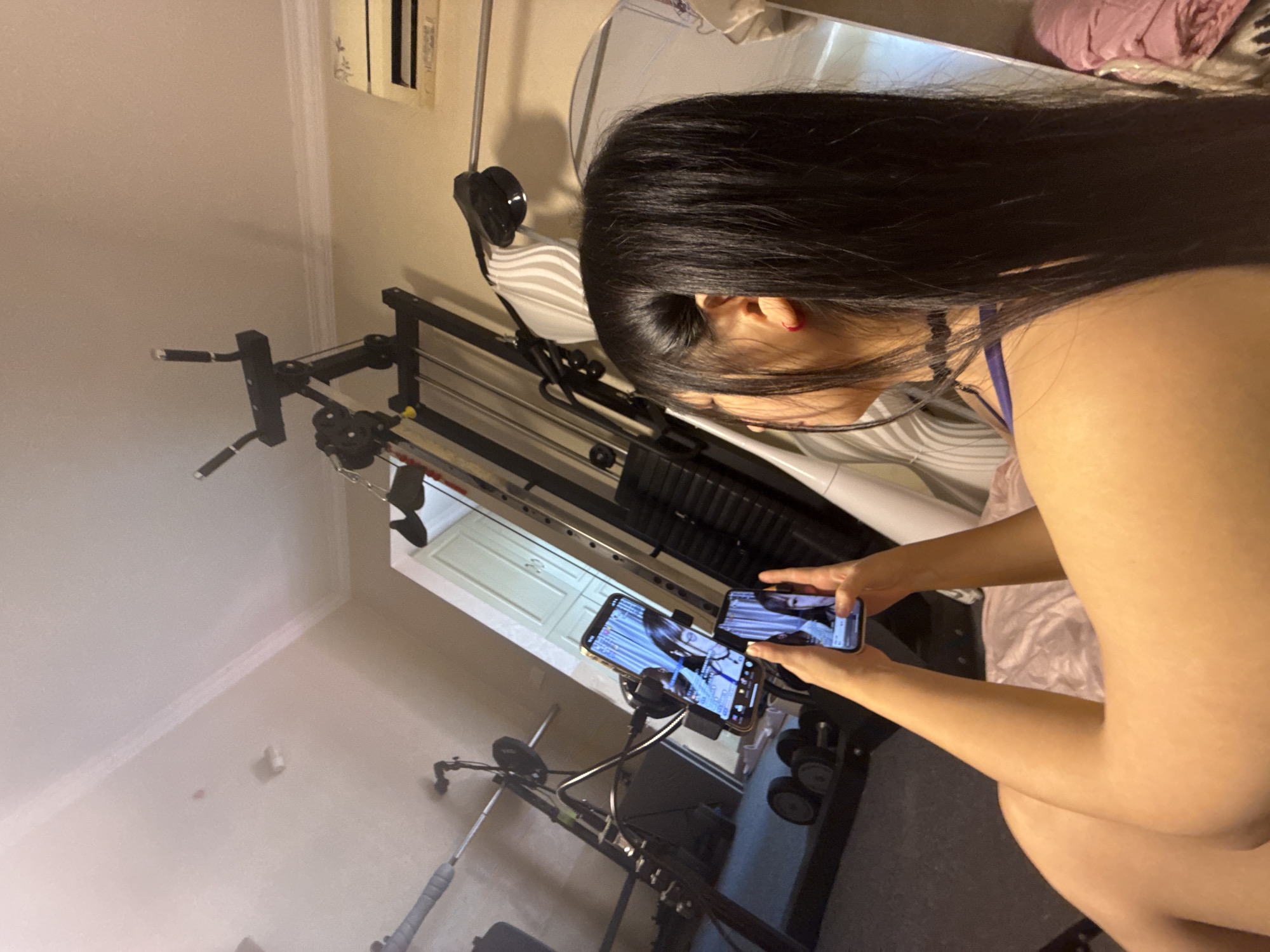 A woman with long, dark hair is holding two cell phones in her hands, seemingly watching a video or interacting with something on the screens. She is positioned in a room that appears to be a home gym, with weightlifting equipment and dumbbells visible in the background. A large mirror is also present, reflecting a portion of the room and a wall with what looks like an air conditioning unit. The overall lighting suggests an indoor setting, possibly during the day given the ambient light. The location is not specified in the image or provided crops.