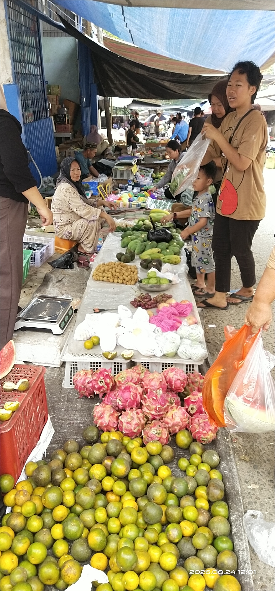 A street market scene unfolds in Tamiang Layang, Indonesia, under a makeshift canopy of blue and black tarps. The foreground is dominated by an abundance of fresh fruit, including piles of dragon fruit, oranges, and what appear to be rambutan or longan. To the left, a red plastic crate holds a sliced watermelon and some halved limes. A digital scale rests on a white tarp. In the mid-ground, several individuals are engaged in commerce. A woman wearing a hijab is seated, seemingly attending to customers or goods. Another woman, in a brown t-shirt, holds a plastic bag containing leafy greens, interacting with a young boy in a patterned shirt. Behind them, more vendors and shoppers are visible, with stalls displaying various items. The overall environment suggests a bustling local market with a focus on fresh produce.