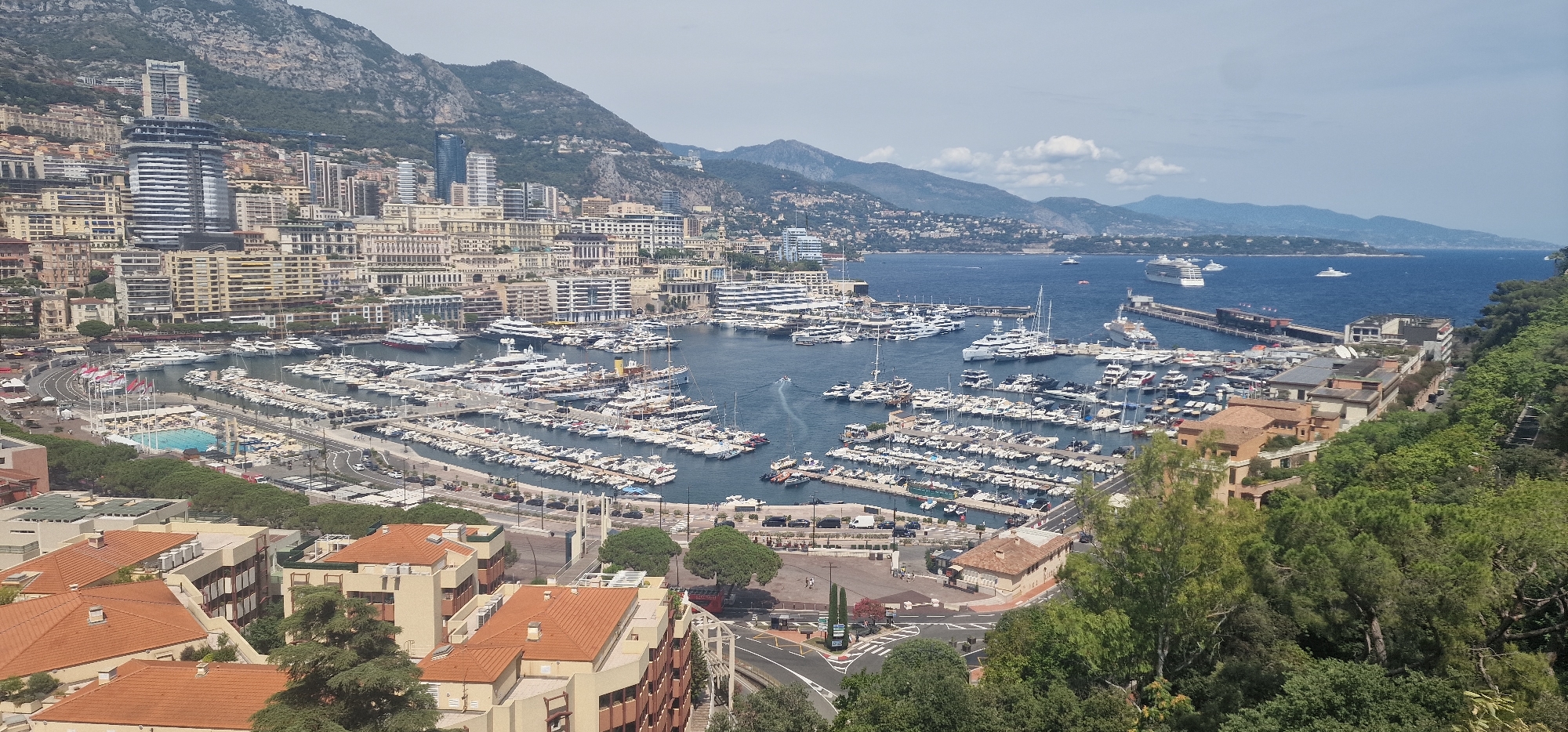 This image depicts a panoramic view of the Port Hercules in Monaco, a principality on the French Riviera. The harbor is filled with a multitude of luxury yachts and smaller boats docked along the piers. In the background, the city of Monaco rises up the steep hillside, characterized by its dense urban landscape of modern high-rise buildings and older, pastel-colored structures. A prominent, sleek, modern skyscraper stands out amongst the other buildings. Beyond the city, a range of mountains forms a dramatic backdrop, their slopes covered in vegetation and dotted with scattered settlements. The blue sea stretches out to the horizon, where a large cruise ship and several other vessels are visible. A swimming pool complex is situated near the waterfront, alongside roads with some car traffic and pedestrians. The foreground is dominated by lush green trees and the terracotta roofs of residential buildings, indicating a vantage point from a higher elevation. The sky is partly cloudy, with patches of blue visible. The overall impression is one of a vibrant and affluent coastal city.