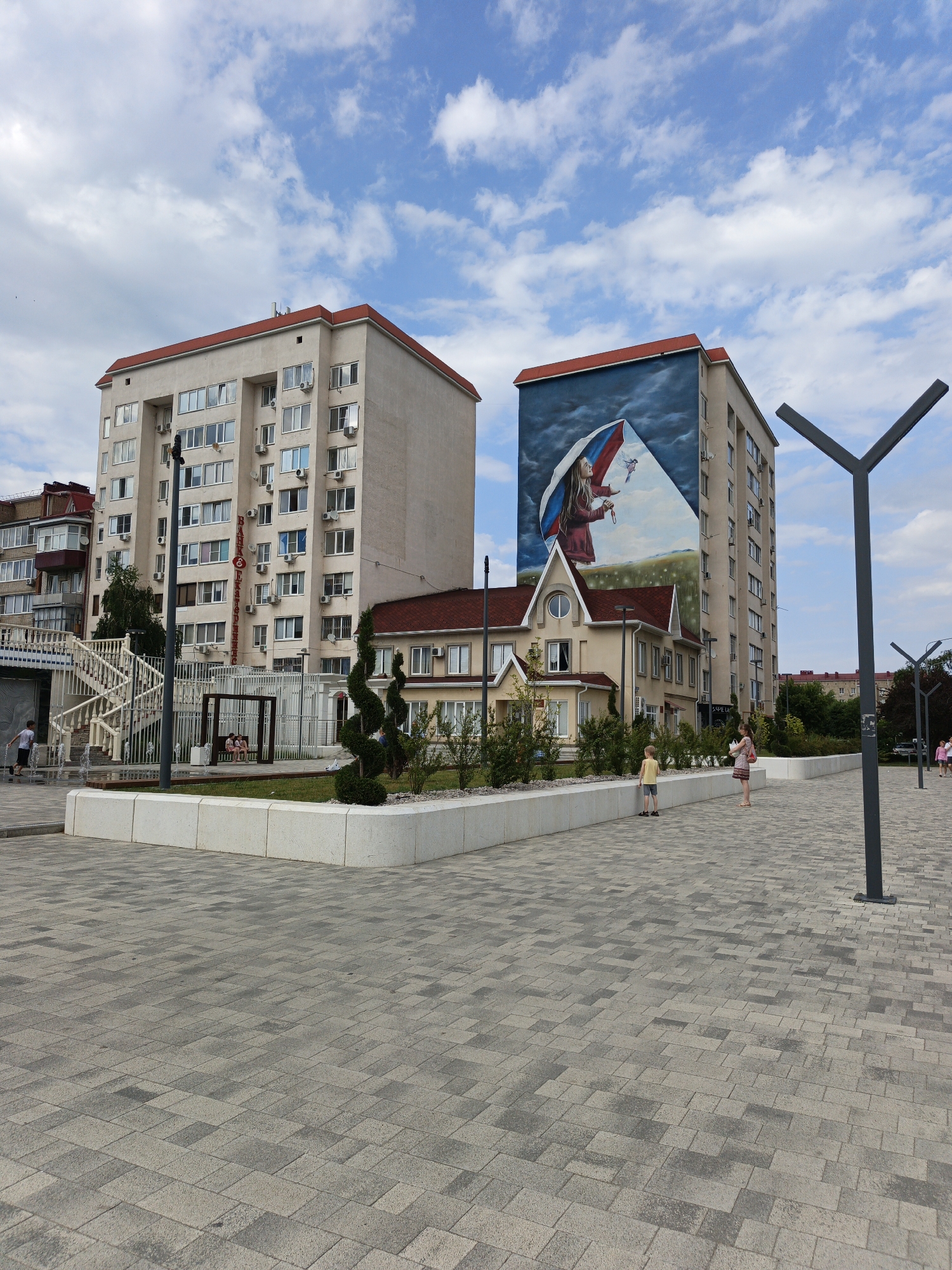 This is a full shot of a modern urban plaza in Goryachiy Klyuch, Russia. The scene is dominated by two multi-story apartment buildings, one with a large mural on its side depicting a young girl holding an umbrella adorned with the Russian flag. The mural is set against a backdrop of a stormy sky and a field. Adjacent to the apartment buildings is a smaller, traditional-style yellow building with a red roof.
The plaza itself is paved with grey interlocking stones, and features a low, white stone planter box filled with green grass and small, sculpted trees. Several people are visible in the plaza. A young boy in a yellow shirt stands near the planter box, while a woman in a patterned dress stands a short distance away. Further down the plaza, two women are walking away from the camera. On the left side of the image, a white staircase with ornate railings leads up to an entrance of one of the buildings. A young boy is running and playing in a water fountain near the base of the stairs.
Streetlights with a modern, Y-shaped design are positioned throughout the plaza. The sky is blue with scattered white clouds, suggesting it is daytime and the weather is pleasant. The overall mood is one of a public space designed for leisure and community gathering. A sign with red lettering is visible on the side of the taller apartment building, but the text is too small to be read clearly.