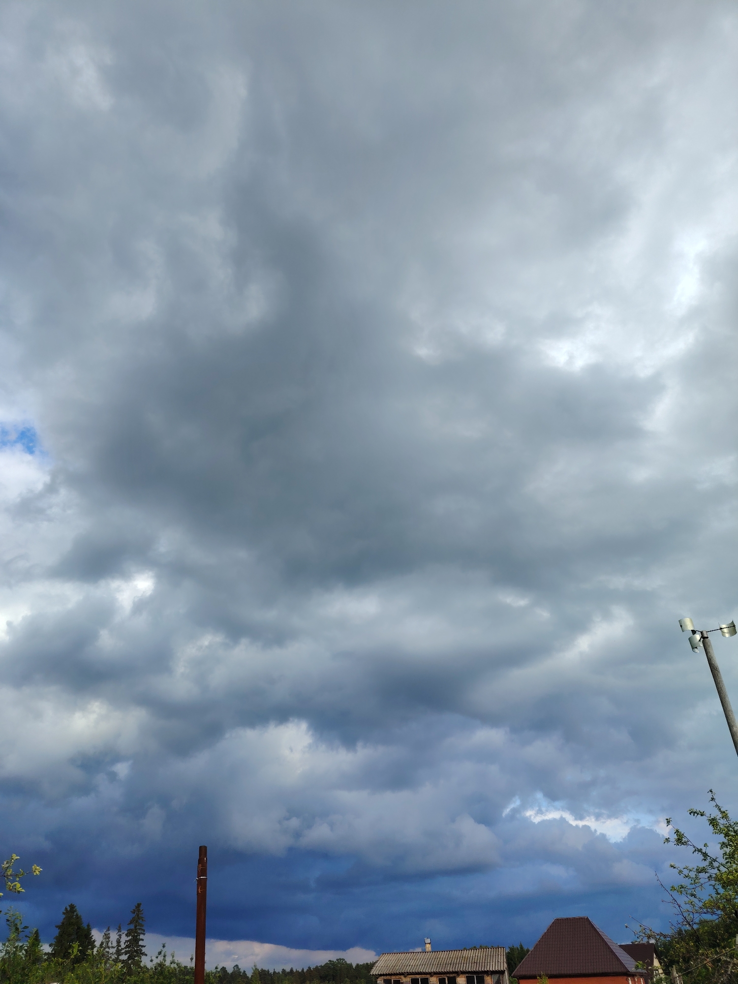 The image depicts a cloudy sky over a location in Chaikovsky, Russia. Below the clouds are visible treetops and sections of buildings with red/brown and light gray roofs. A vertical post is in the lower left of the frame, and a light pole is on the right. The sky dominates the frame with varying shades of gray clouds.