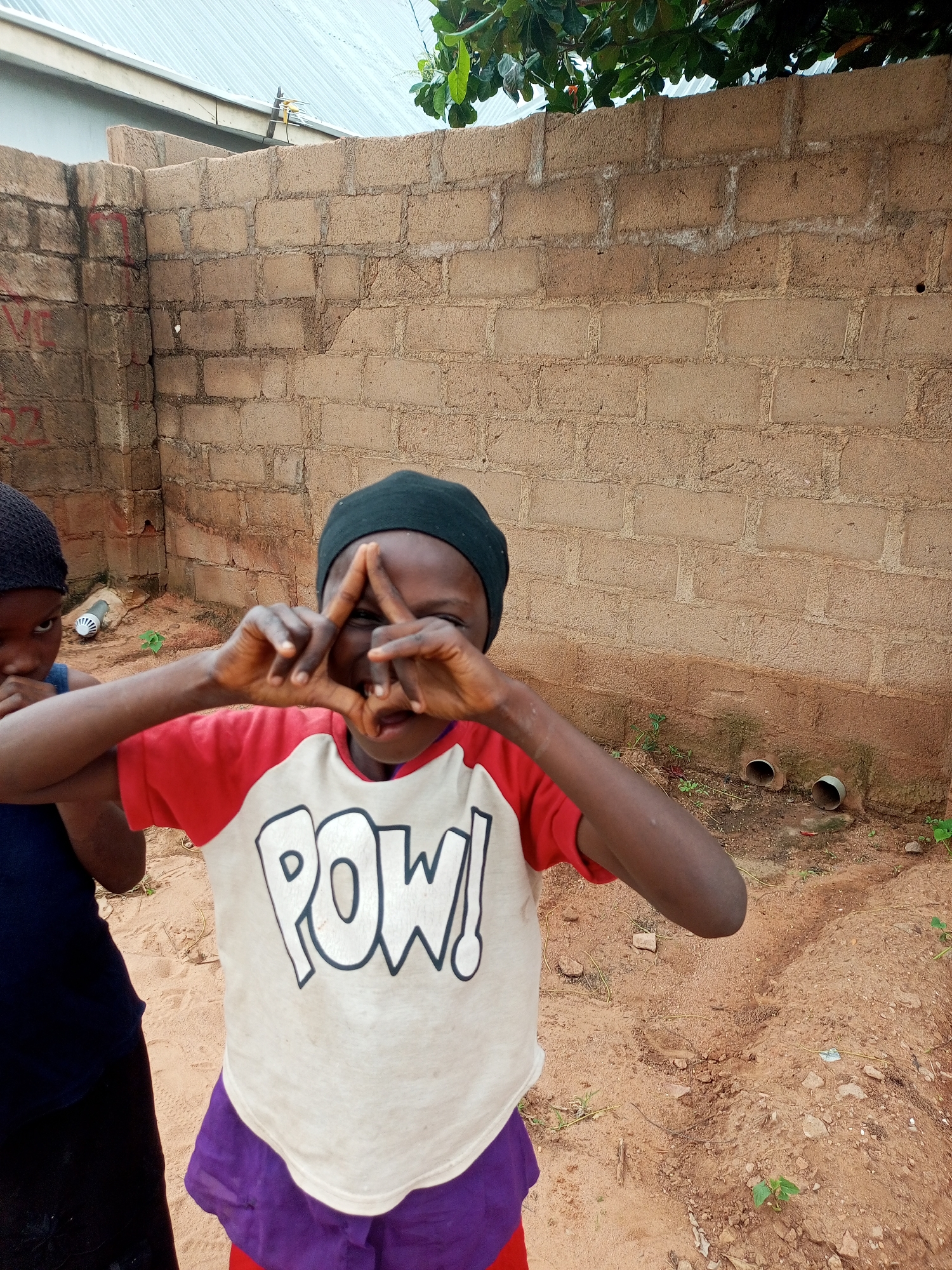 The image depicts two children in an outdoor, dusty environment in Tempure, Nigeria. The child in the foreground, wearing a red and white t-shirt with "POW!" printed on it and a black beanie, is making a hand gesture that forms a triangle over their face, smiling at the camera. To the left, another child in a dark blue top and black head covering is partially visible, looking towards the camera. The background features a rough, brick wall and a sandy ground with some scattered rocks and small green plants. Two white pipes protrude from the wall near the ground. The overall scene suggests a casual, everyday setting in a residential area.
