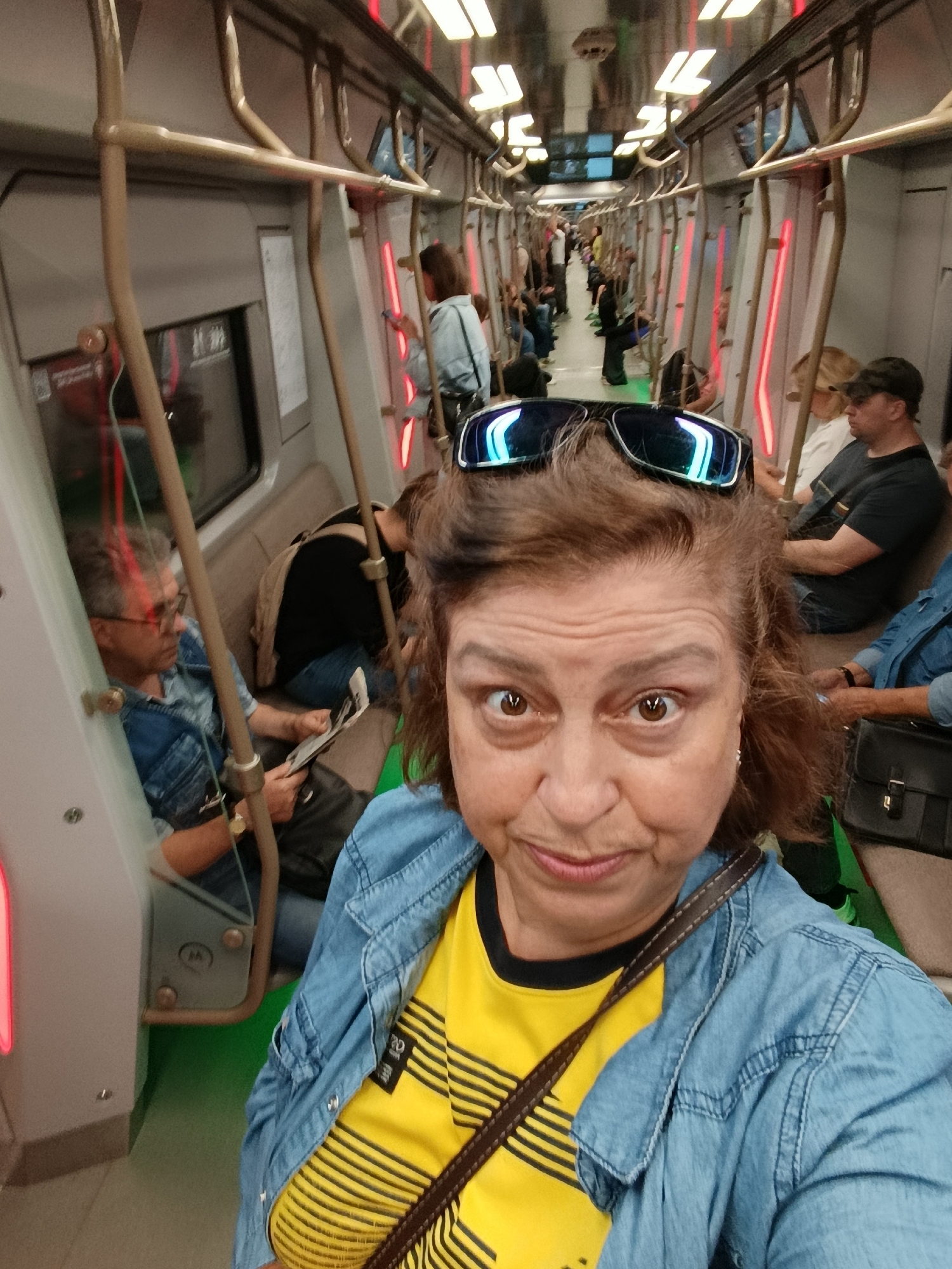 This is a selfie taken by a woman with medium-length brown hair, wearing sunglasses on top of her head and a denim jacket over a yellow and black striped shirt. She is in a subway car, with other passengers visible in the background. To her left, a man in a denim shirt is reading a newspaper. Further down the car, more passengers are seated and standing. The interior of the subway car features overhead handrails, red and white lighting, and beige seats. The scene appears to be in the city of Unknown, Unknown.
