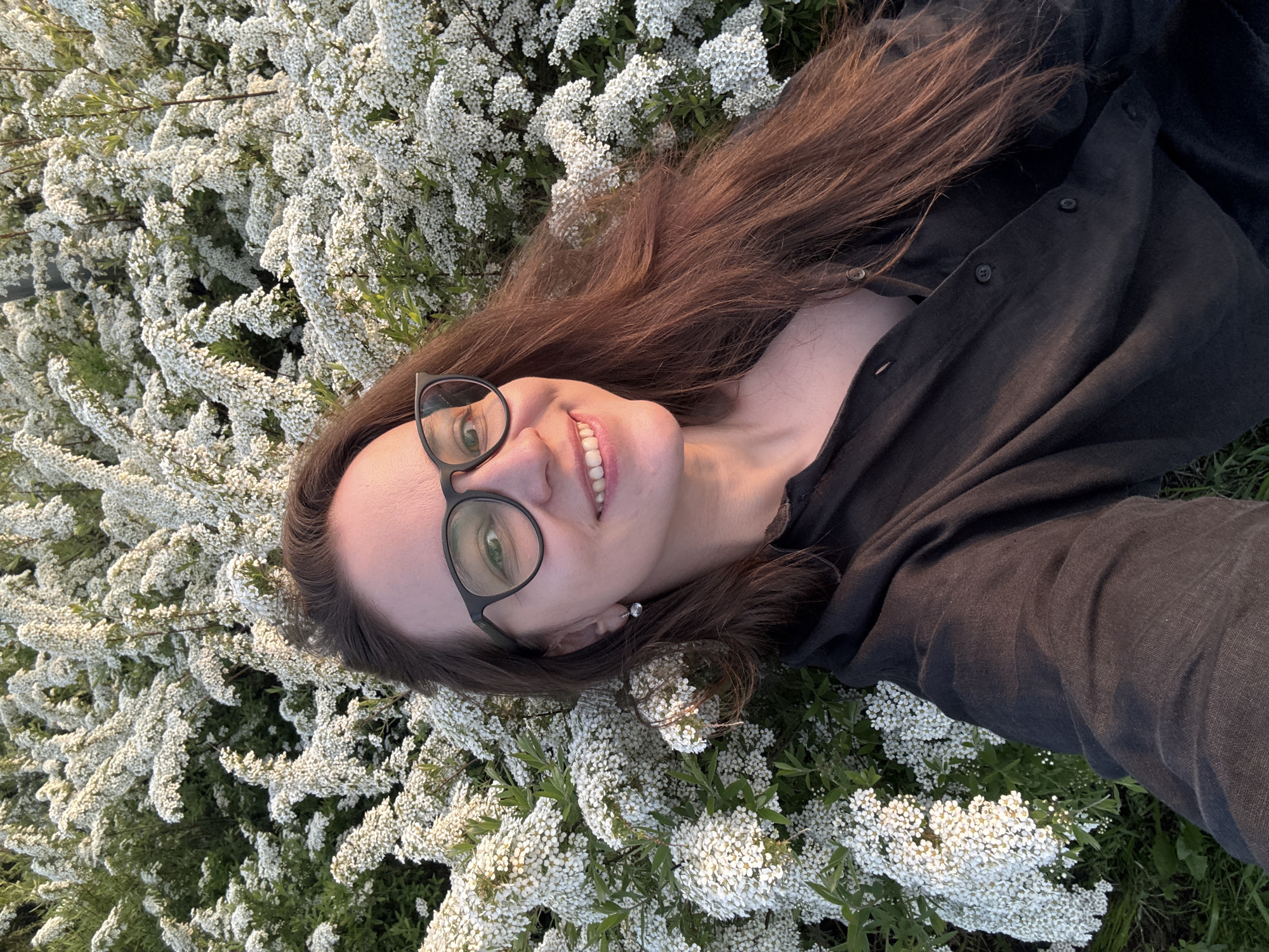 A woman with long brown hair and glasses lies in a field of small white flowers, possibly spirea, wearing a dark colored button-down shirt. She smiles at the camera. The location appears to be outdoors, possibly a park or field in the city of Nizhny Novgorod, Russia.