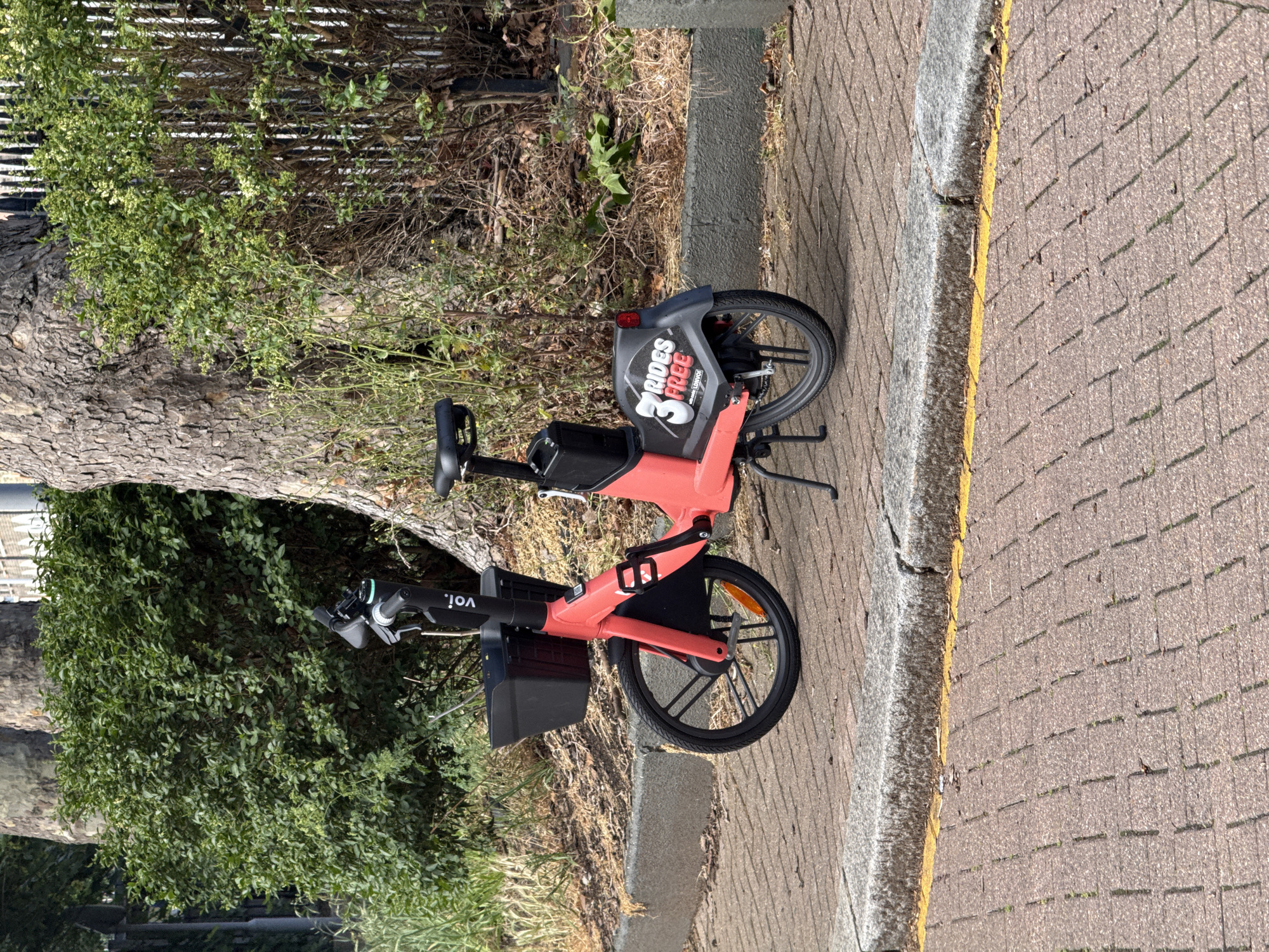 A coral-colored Voi e-bike is parked on a block-paved sidewalk during daylight. The bike, positioned on its kickstand, features a black front basket and a rear mudguard. To the left of the bike, a large tree with textured bark stands alongside dense green foliage and bushes. A dark metal fence is partially visible behind the greenery. To the right, the paved sidewalk continues, bordered by a grey concrete curb with a weathered yellow line painted along its edge. Bright sunlight casts clear shadows on the pavement, indicating a sunny day.
No people are visible in the scene; the e-bike is stationary.
Notable details include the bike's prominent coral color and black components. The rear mudguard displays a logo with the text "3 RIDES FREE" in white and red, with "VOI" written below it in smaller letters. The handlebars or stem also show the "voi." logo, which appears upside down due to the image's orientation. The general setting suggests an urban or semi-urban environment, consistent with the provided location context of London, United Kingdom.