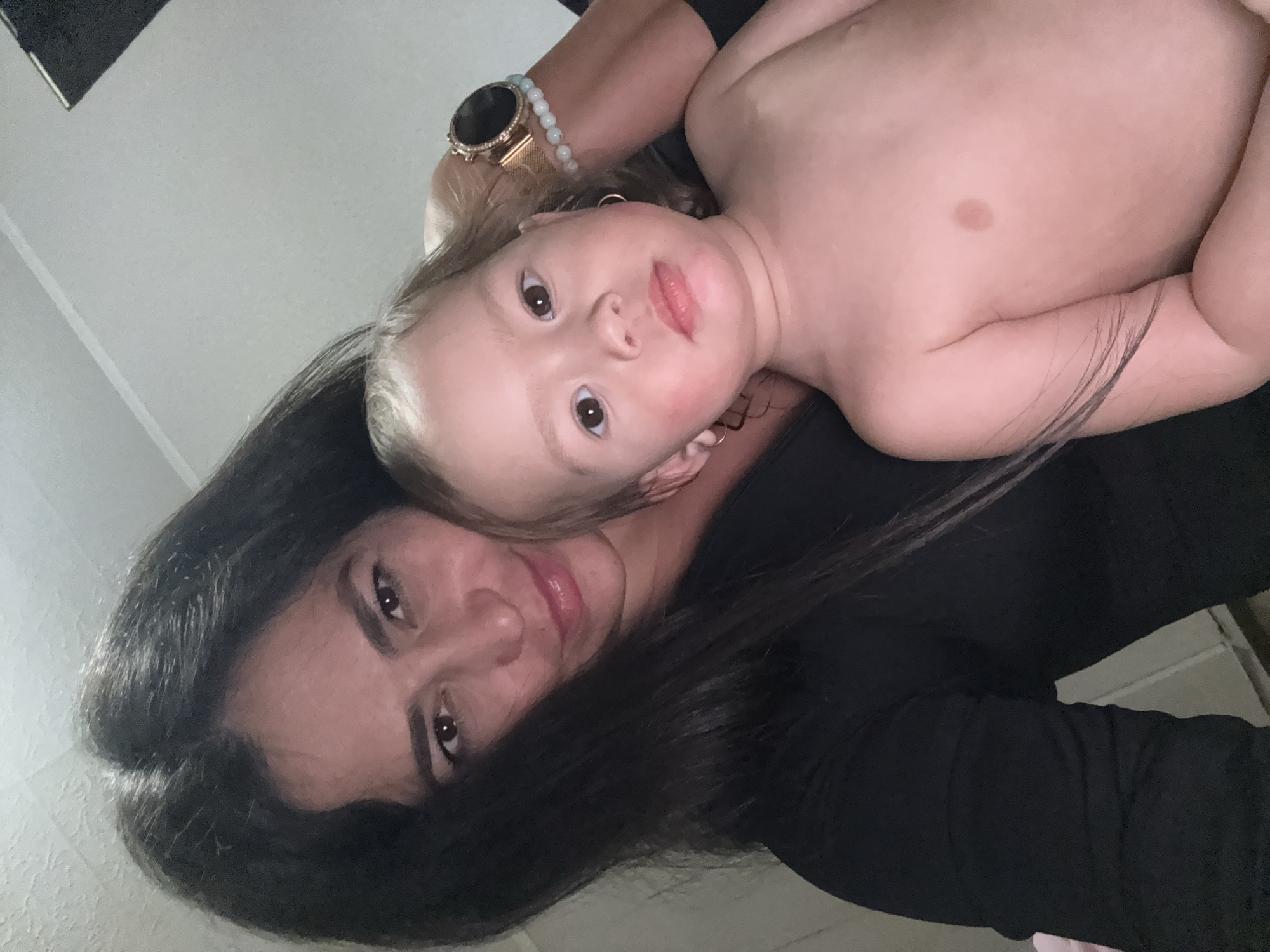 This close-up, rotated selfie features an adult woman and a baby or toddler, captured indoors in Chaikovsky, Russia. The perspective is from above, looking down on the subjects.
In the lower and central part of the frame, a woman with long, dark hair and dark eyes looks directly at the camera with a calm expression. She is wearing a dark, long-sleeved top. On her left wrist, a gold-toned watch with a mesh band and a light blue beaded bracelet are visible.
Cradled next to the woman, in the upper portion of the image, is a young child. The child has light-colored hair and dark, attentive eyes, also looking directly into the camera. The child appears to be shirtless, revealing a fair-skinned torso, and has a distinct brown birthmark or mole on the upper chest area. Small hoop earrings are visible in the child's ears. Some of the woman's dark hair falls gently over the child's body.
The background consists of a simple, light-colored wall or ceiling, suggesting an uncluttered indoor environment. The lighting is soft and even, characteristic of an indoor setting. The image conveys an intimate, tender moment shared between the woman and the child. No text is visible in the media file.