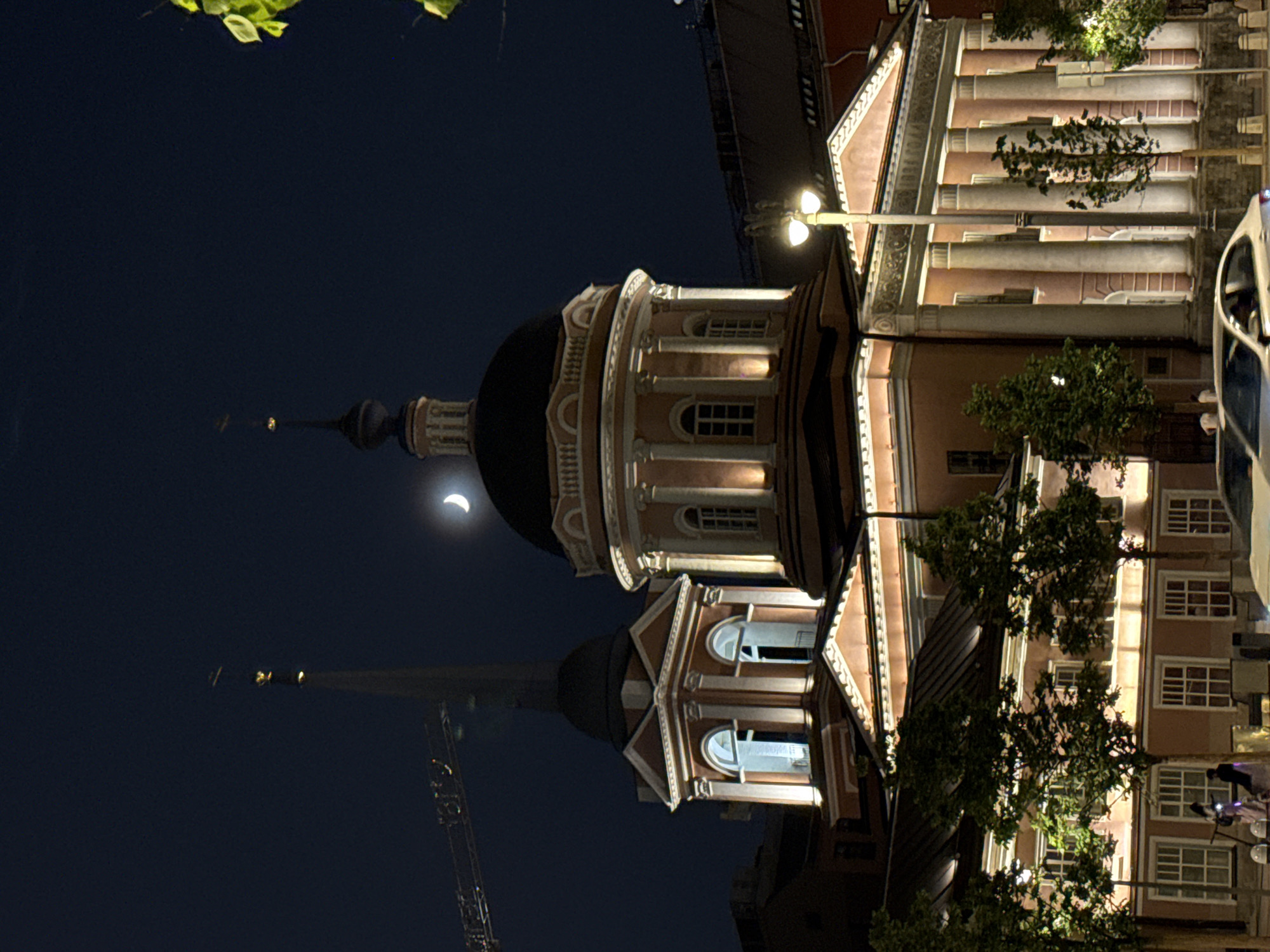 The image captures a nighttime scene in Moscow, Russia, featuring a reflection of the church in a body of water. The primary focus is on the architecture of the building, which includes a black dome and an ornate, tiered structure with windows. A crescent moon is visible in the upper left part of the image. The scene appears to be artificially illuminated, with streetlights providing light on the building and its reflection. A crane is barely visible in the background. A vehicle is visible on the right.