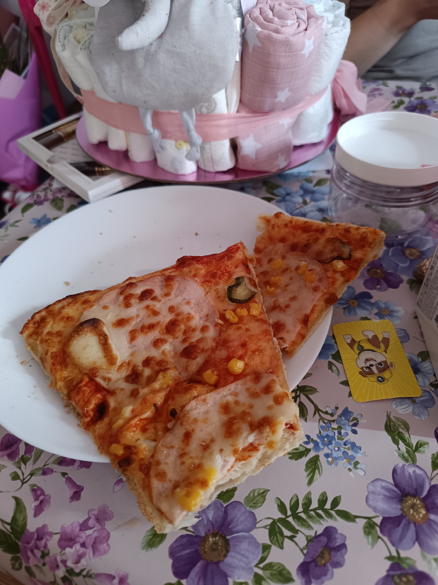 The image shows two slices of pizza on a white plate, placed on a floral tablecloth. The pizza appears to have cheese, ham, corn, and jalapenos as toppings. Beside the plate is a yellow card with a cartoon character on it, along with a clear plastic jar with a white lid. In the background, there's a diaper cake with a plush elephant on top, and a pink bag. The location is likely indoors, possibly a home, with the floral tablecloth adding a domestic touch. There is no clear indication of being in the city of Sofia, Bulgaria.
