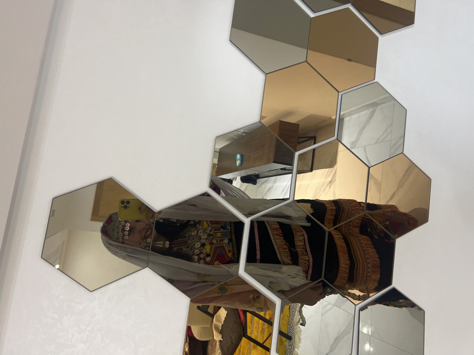 A collection of gold and mirrored hexagonal tiles adorns a white wall, reflecting a room interior. A woman in traditional attire is seen in the reflection, taking a selfie with a phone. The room features a wooden desk, a chair, and a hint of yellow and red fabric. The mosaic creates a fragmented view of the scene, blending the room's elements with the woman's self-portrait.