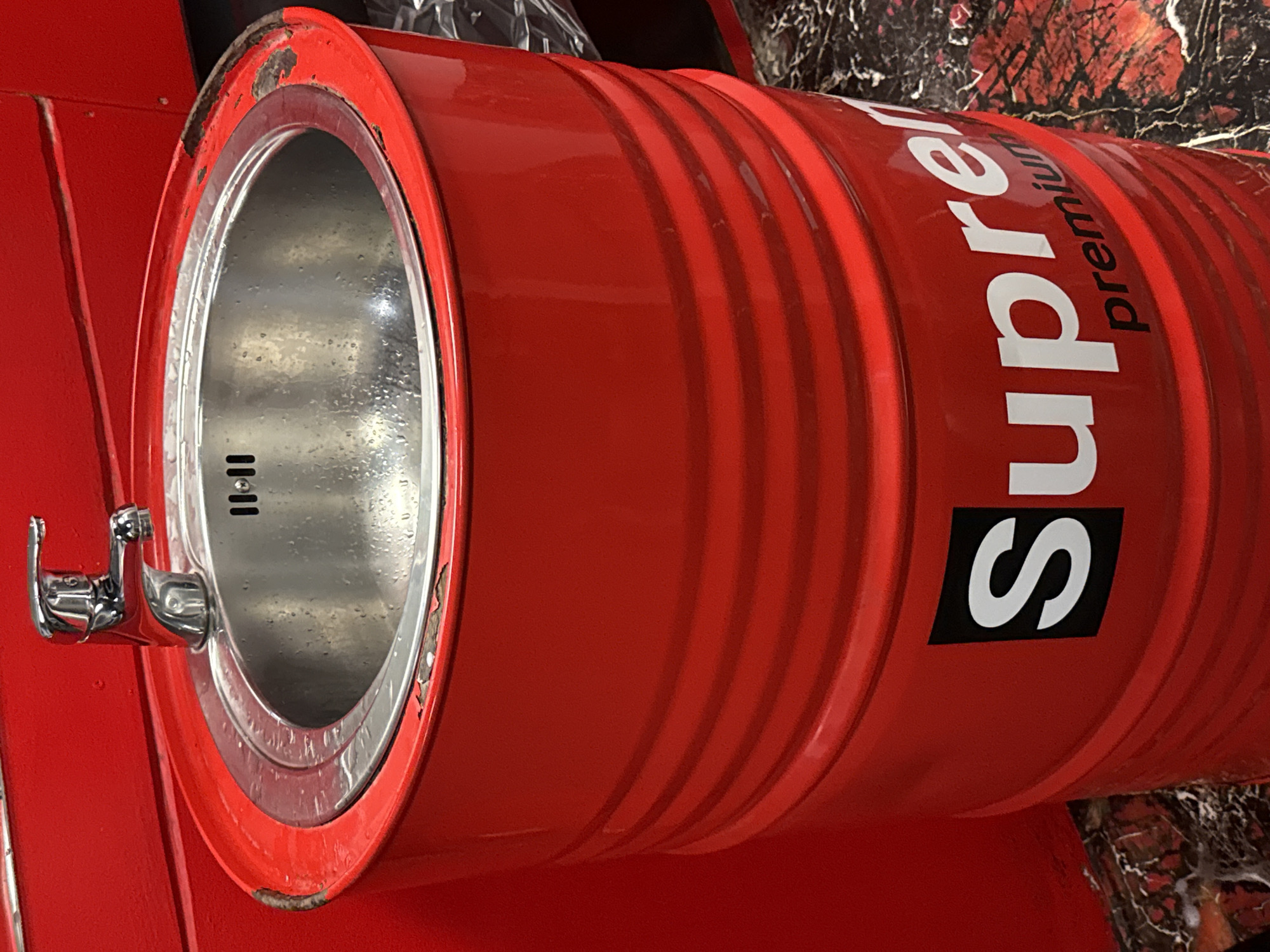 A close-up shot shows a red metal barrel repurposed as a sink. The barrel is emblazoned with the word "Supreme" in a vertical orientation. The sink has a metal faucet and a drain. The barrel and sink are located in Sofia, Bulgaria.