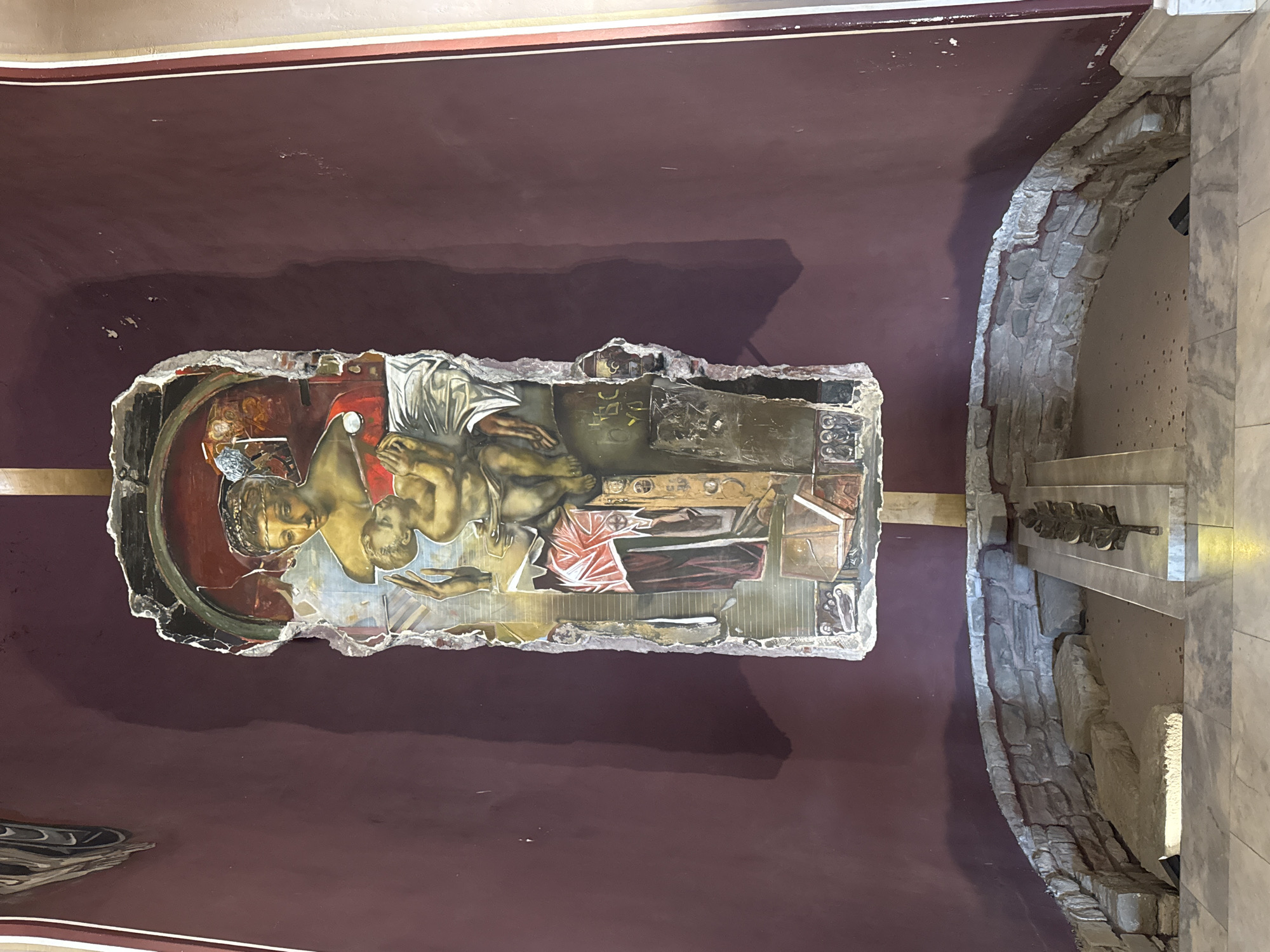 The image shows a damaged fresco depicting the Virgin Mary holding the infant Jesus. The fresco is mounted on a wall in what appears to be a historical or religious site. Below the fresco, there is a stone enclosure with a metal sculpture resembling a laurel wreath placed on a marble slab. The surrounding walls are painted a deep red, and the floor is tiled. The fresco itself is fragmented and weathered, with visible cracks and missing sections. The figures in the fresco are rendered in a traditional Byzantine style. The overall impression is one of age and historical significance, suggesting the site is a place of worship or a preserved historical monument. This site is located in the Church of the Forty Holy Martyrs in Veliko Tarnovo, Bulgaria, which houses relics of the saint and is a significant historical monument.