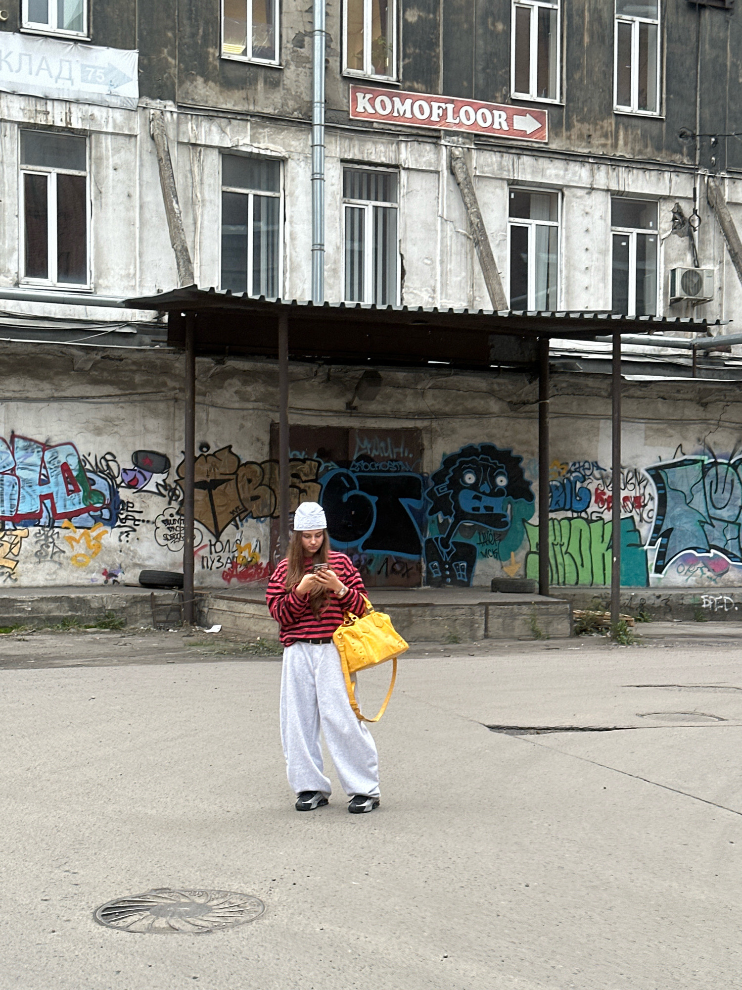 The image depicts a young woman standing on an asphalt street in front of a graffiti-covered concrete wall and an aged, dilapidated building. The woman is wearing a white beanie, a red and black striped long-sleeved shirt, and light-colored baggy pants. She is holding a smartphone in her hands and has a bright yellow handbag slung over her shoulder. The building behind her has multiple windows and appears to be a multi-story structure, with a faded sign that reads "KOMOFLOOR" with an arrow pointing to the right. The wall in the foreground is extensively covered in vibrant graffiti art. The street itself shows signs of wear, with a manhole cover visible in the lower left quadrant of the image. The overall atmosphere is urban and somewhat gritty. This scene is located in Saint Petersburg, Russia, at an undisclosed address, though the building's architecture and the presence of Russian signage suggest this location.