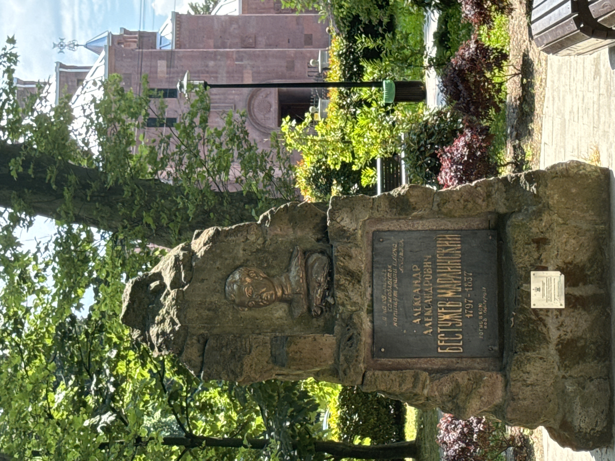 The image shows a monument in a park in Sochi, Russia. The monument is a large stone structure with a bronze bust of a man at the top. Below the bust is a plaque with text in Russian. In the background, there is a church with a red brick facade and a white roof. There are trees and bushes in the park, and a bench and a trash can are visible. The sky is cloudy.